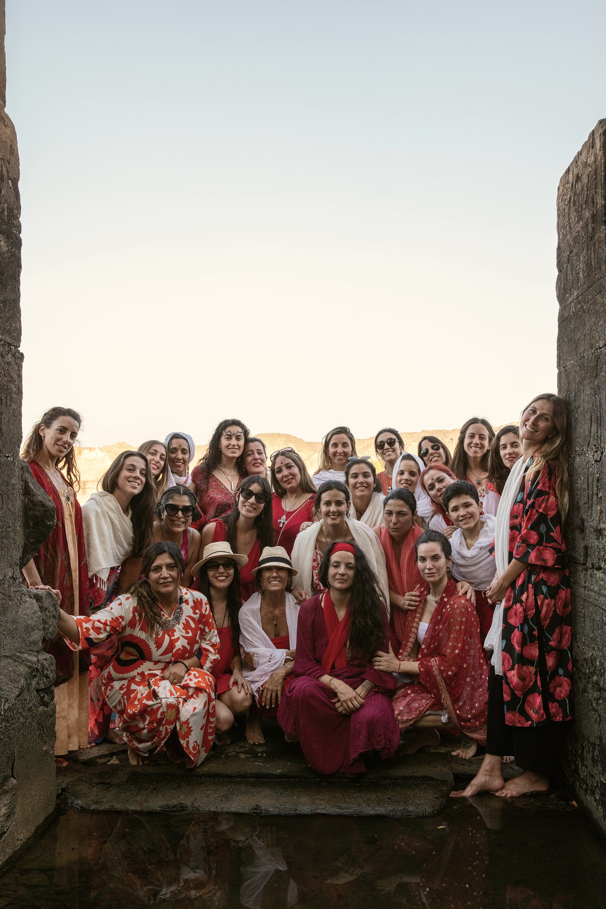 Un grupo de mujeres en colores vibrantes, algunas con sombreros, posando juntas en un entorno al aire libre con paredes de piedra.