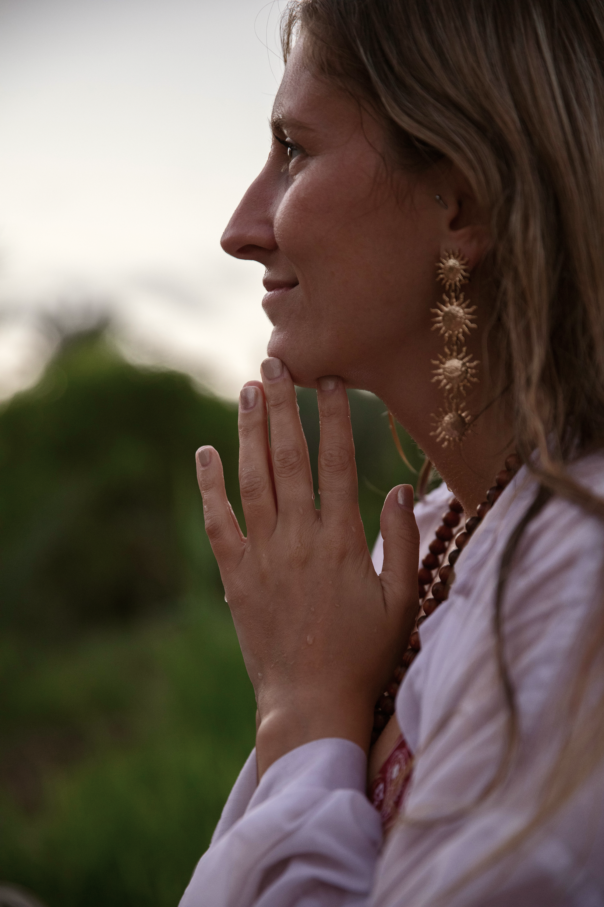 Retrato lateral de una mujer con pendientes grandes y collar de madera, tocada con cabello rubio, en un entorno natural.