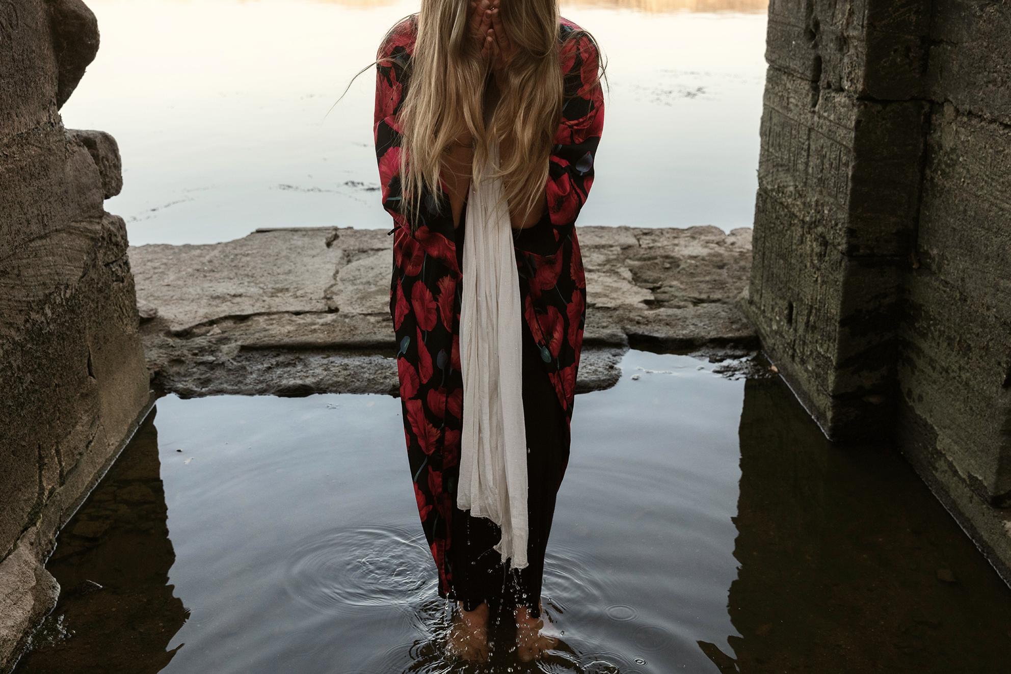 Mujer arrodillada en el agua, cubriéndose la cara con las manos, entre muros de piedra junto a un cuerpo de agua.