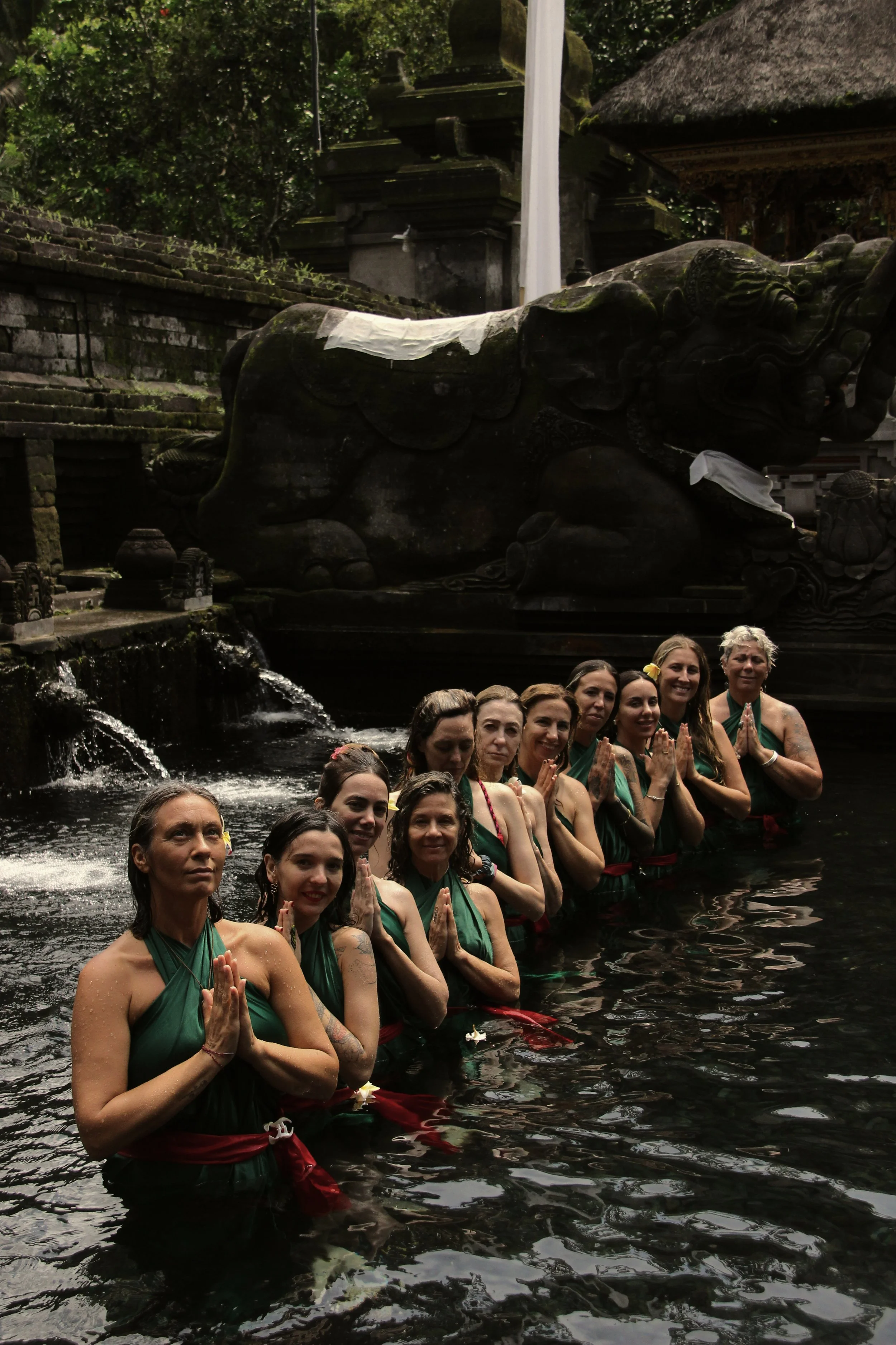 Grupo de personas realizando una ceremonia en un río, con esculturas de animales de piedra en un entorno cultural tradicional de Bali, Indonesia.