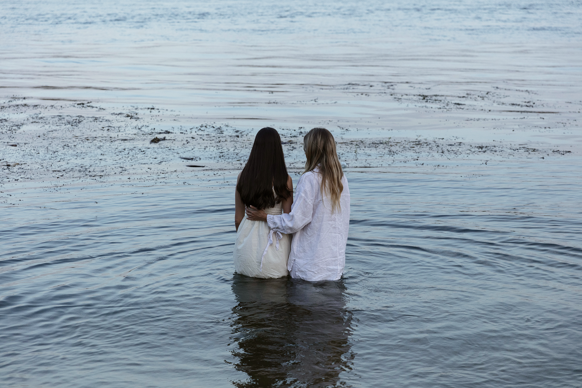Dos mujeres abrazadas, una de cabello negro y otra de cabello rubio, de pie en el agua del mar.