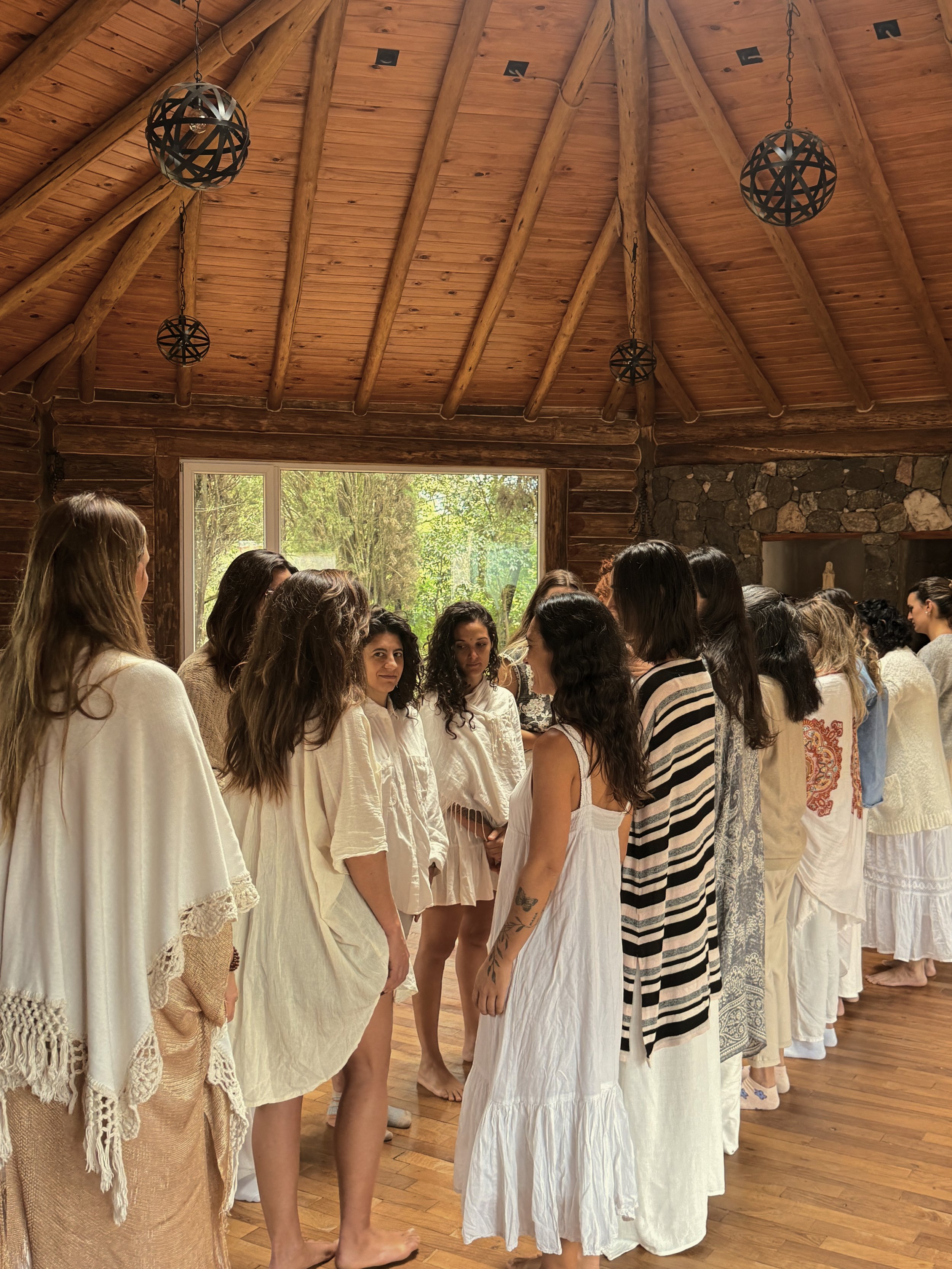 Grupo de mujeres en una ceremonia en una cabaña de madera, vestidas con ropa blanca, algunas con tatuajes, en un ambiente natural.