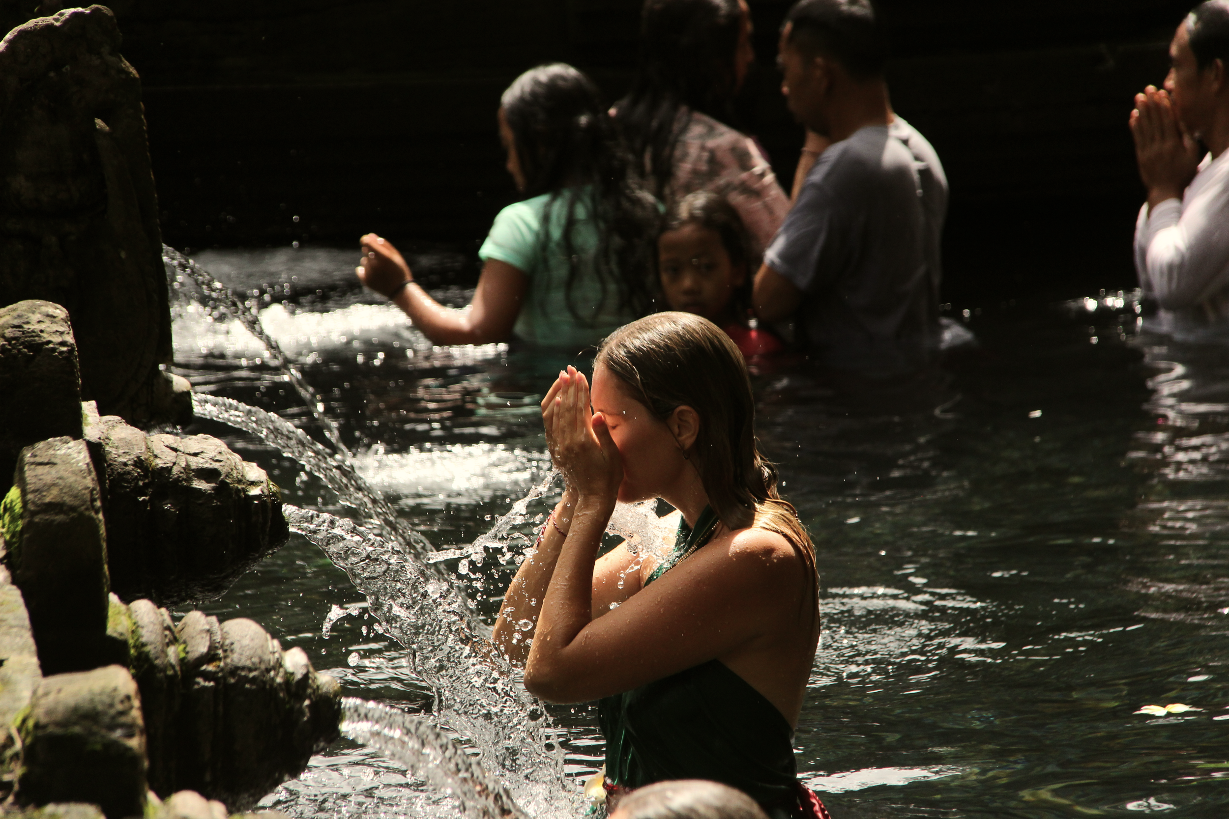 Personas en el agua en un río, varias con las manos juntas en oración y una con las manos en la cara, en un entorno natural