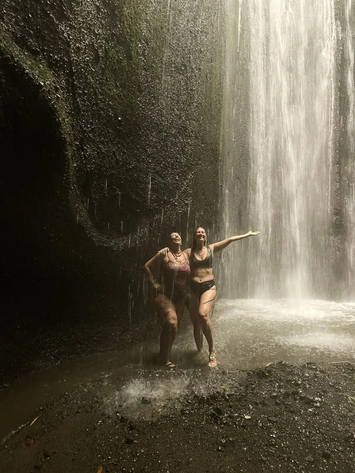 Dos mujeres en bikini bajo una cascada en un entorno natural.