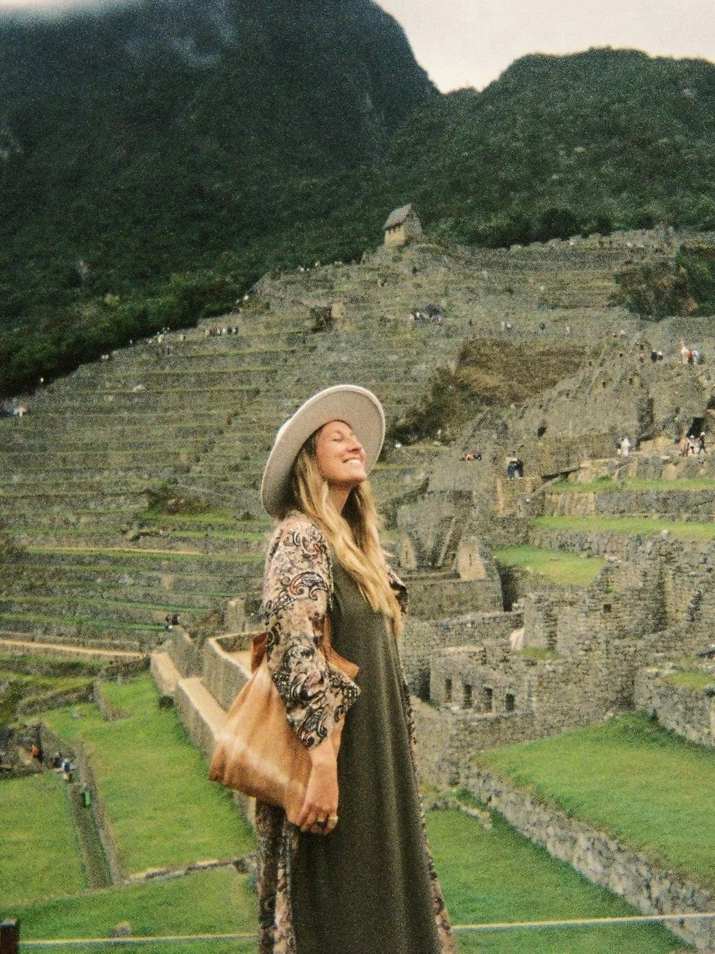 Mujer sonriendo con sombrero grande frente a las ruinas de Machu Picchu en Perú al atardecer.