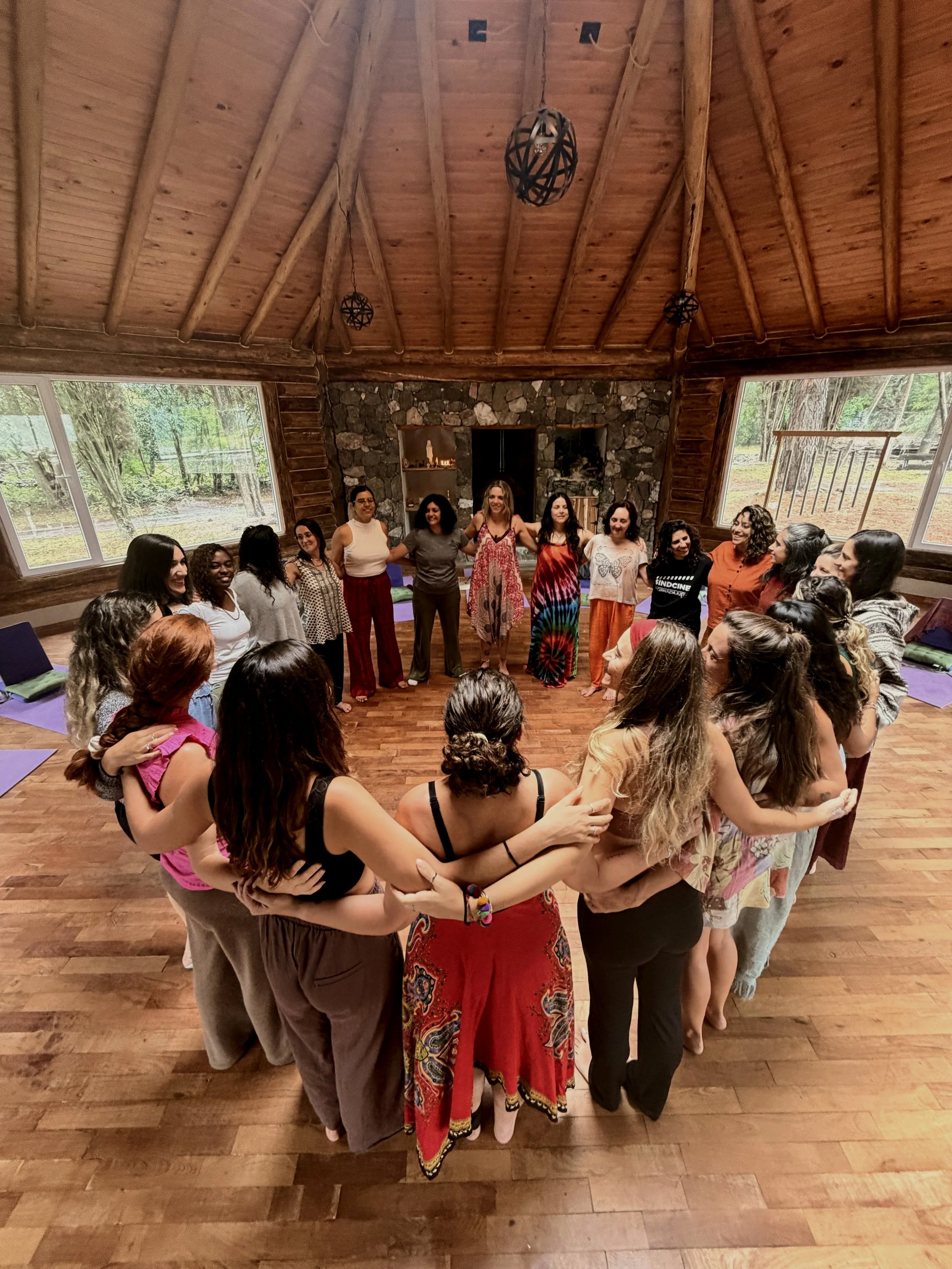 Grupo de mujeres formando un círculo en una sala con paredes de madera y ventanas grandes, en lo que parece ser un taller o reunión.