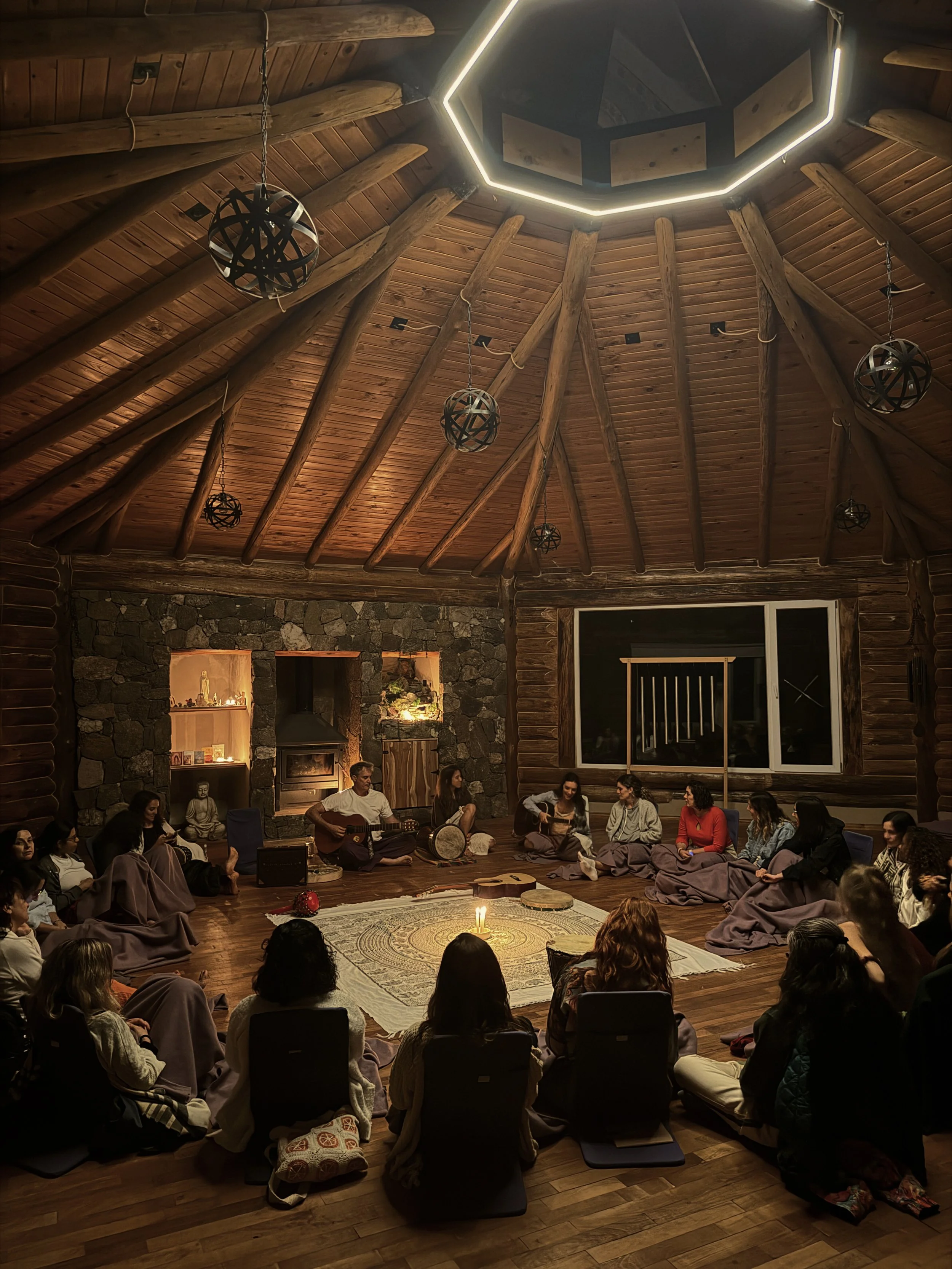 Grupo de personas en un círculo en una sala rústica de madera, participando en una ceremonia o reunión espiritual, con músicos tocando instrumentos tradicionales y luz suave.
