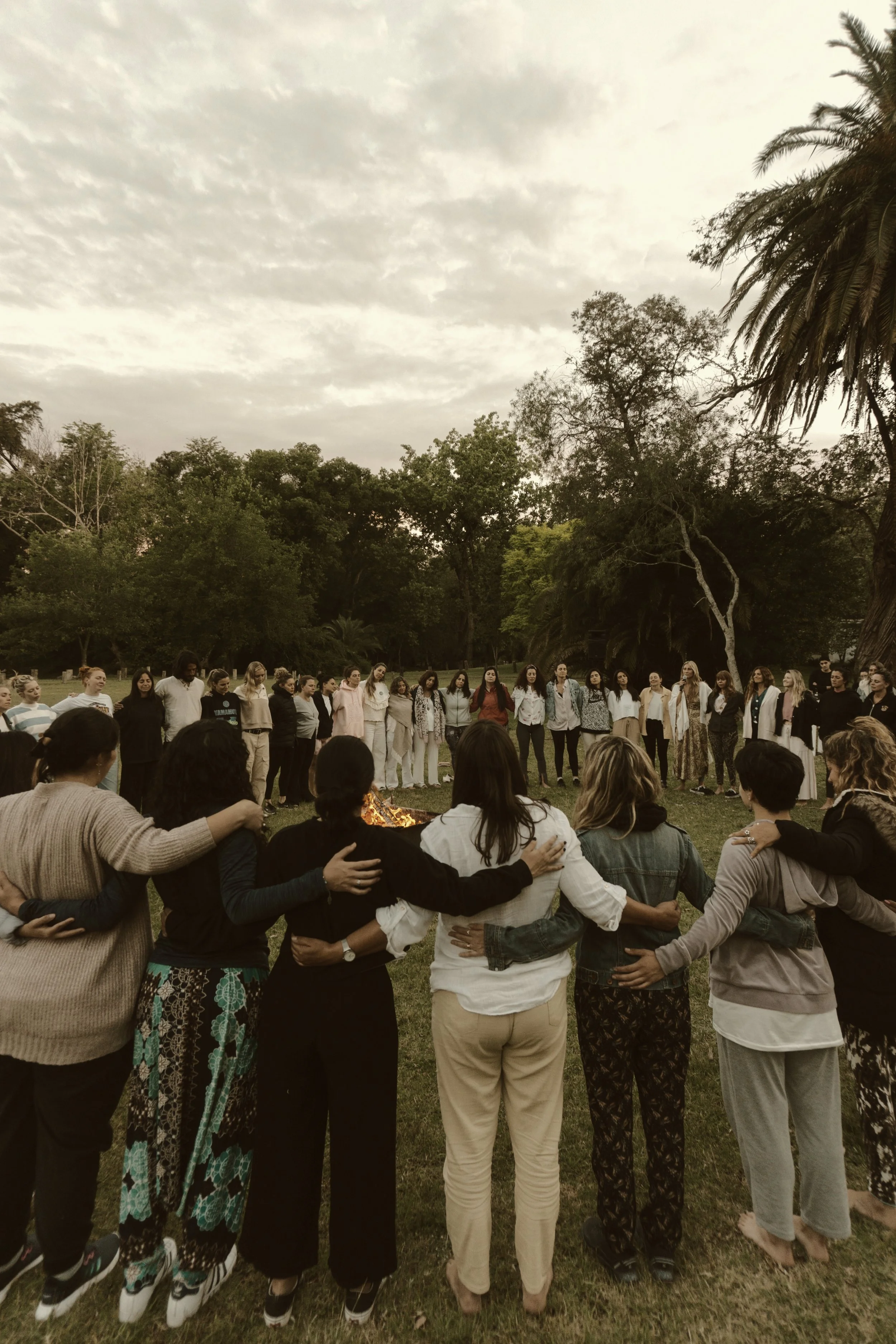 Grupo de personas de diversas edades y géneros formando un círculo, con brazos sobre los hombros de las personas adyacentes, en un espacio al aire libre con árboles verdes y cielo nublado, participando en una ceremonia alrededor de una fogata.