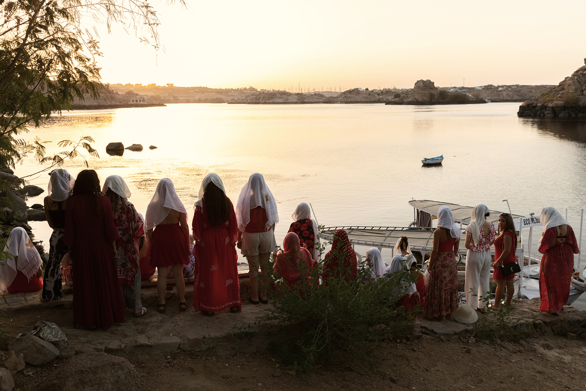 Grupo de personas con pañuelos blancos en la cabeza, reunidas frente a un río al atardecer, algunos sentados y otros de pie, con embarcaciones en el agua y un paisaje de colinas y árboles en el fondo.