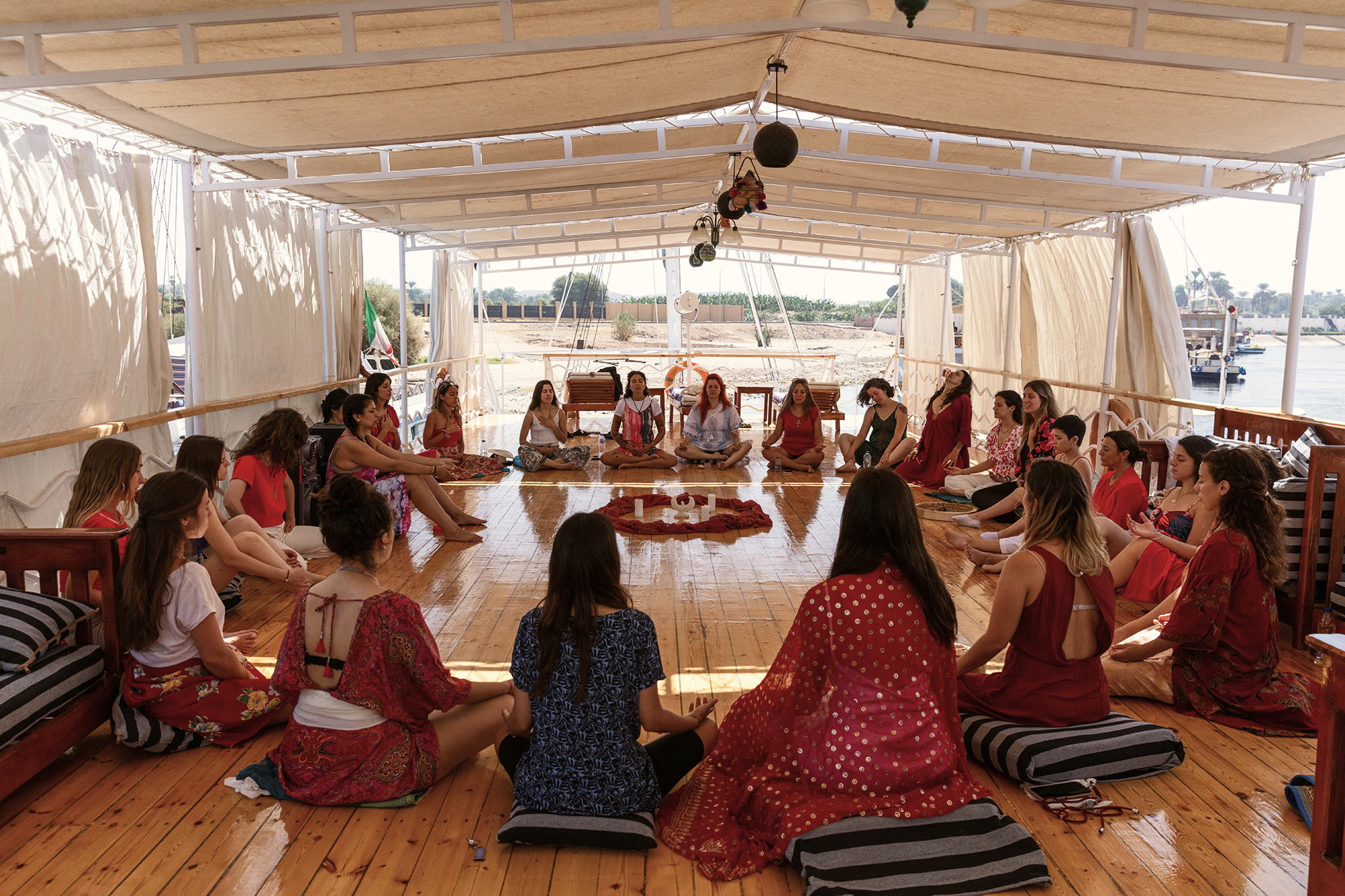 Grupo de mujeres y niños sentados en círculo en una embarcación cubierta, participando en una actividad de meditación o yoga, decorada con velas en el centro.