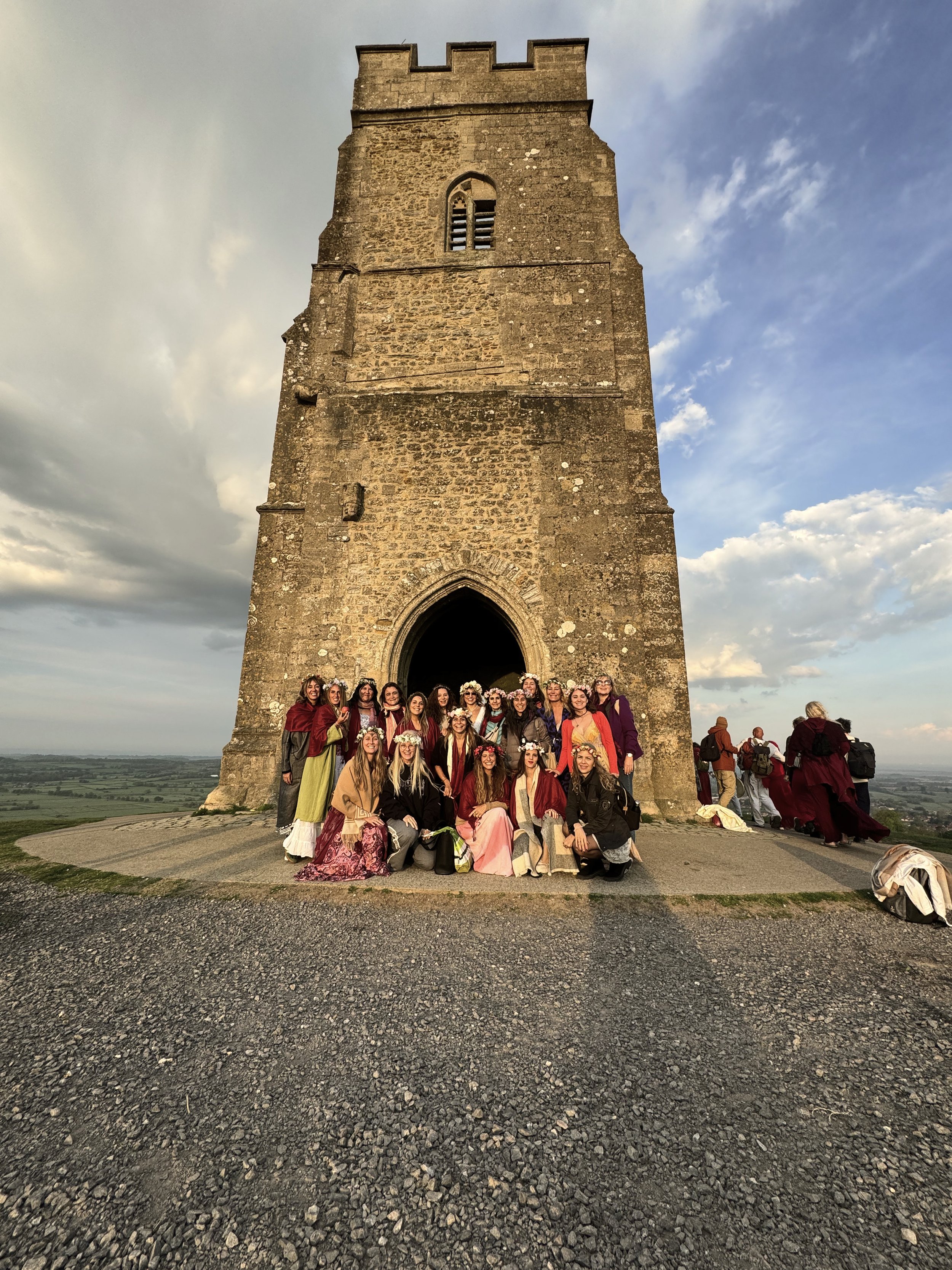Grupo de personas vestidas con ropa y accesorios de estilo bohemio, posando frente a una antigua torre de piedra en un pueblo rural. El cielo está parcialmente nublado y se aprecia un paisaje amplio con campos en el fondo.