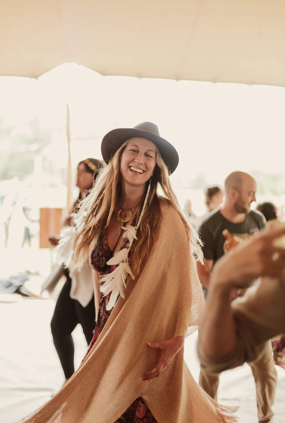 Una mujer sonriendo, vestida con un sombrero, un vestido de colores y una bufanda de flecos, en un evento al aire libre.