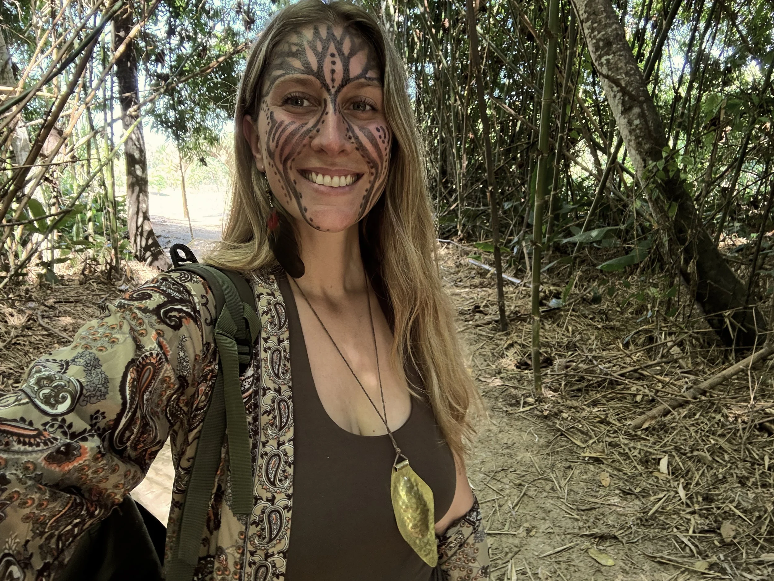 Mujer sonriente con rostro pintado en un bosque, lleva una mochila y un collar de hoja amarilla.