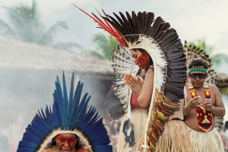 Personas con trajes tradicionales con penachos de plumas en una ceremonia cultural en un entorno natural.