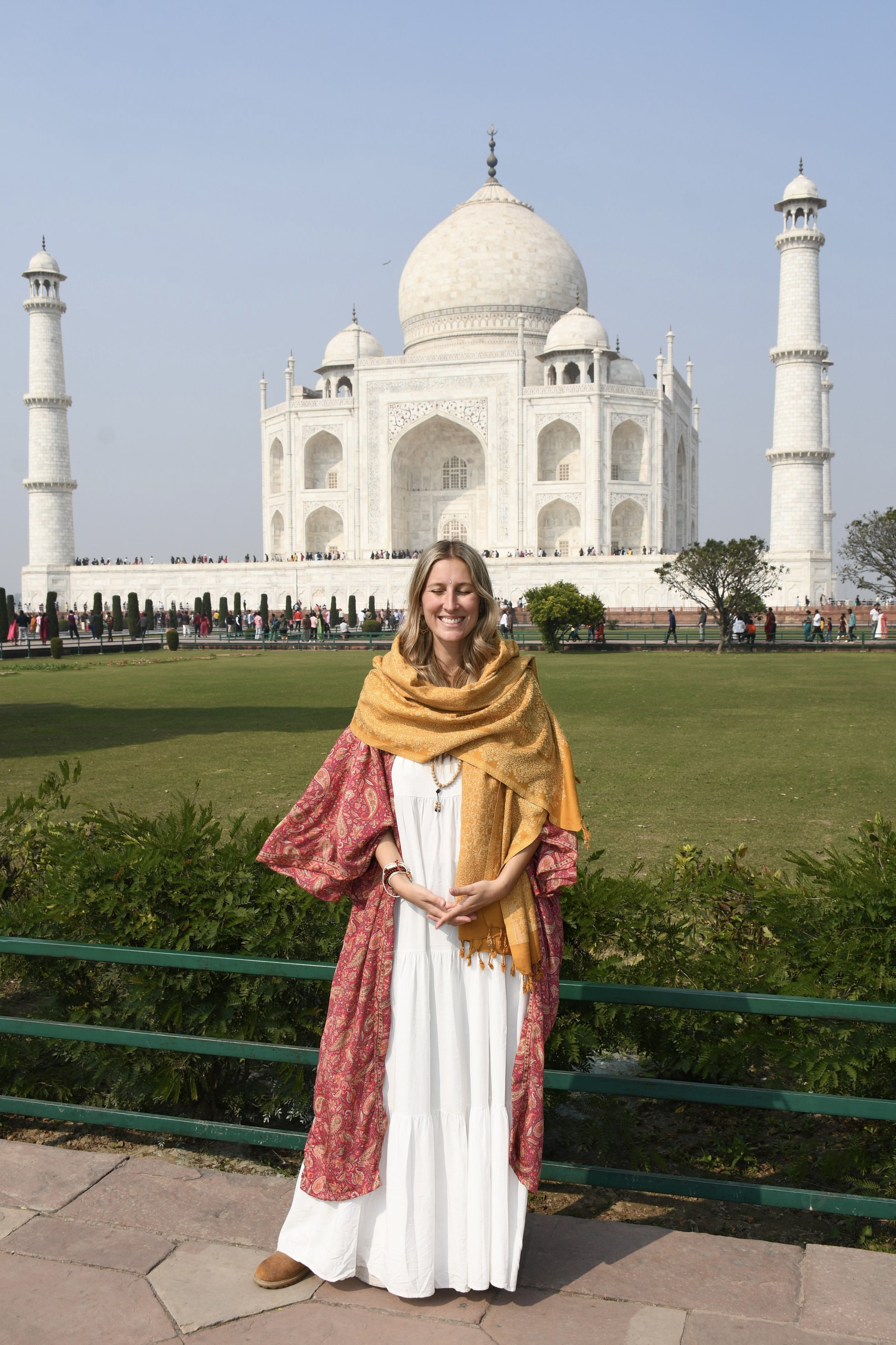 Mujer sonriendo con túnica blanca, bufanda dorada y túnica estampada en primer plano, en frente del Taj Mahal en India.