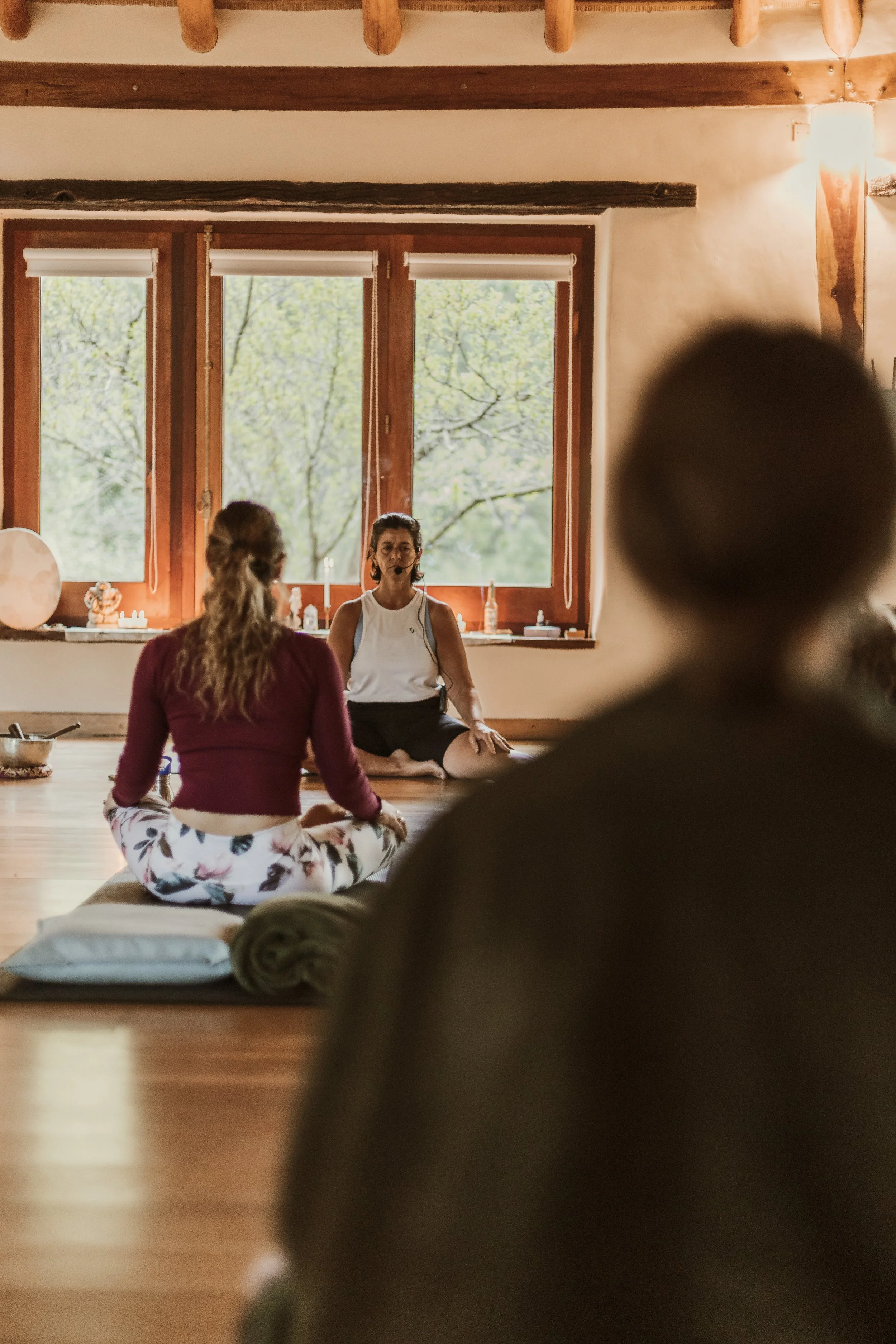 Rutina de yoga en un estudio con ventanas grandes, dos mujeres sentadas en postura de meditación y una instructora con micrófono en el centro.