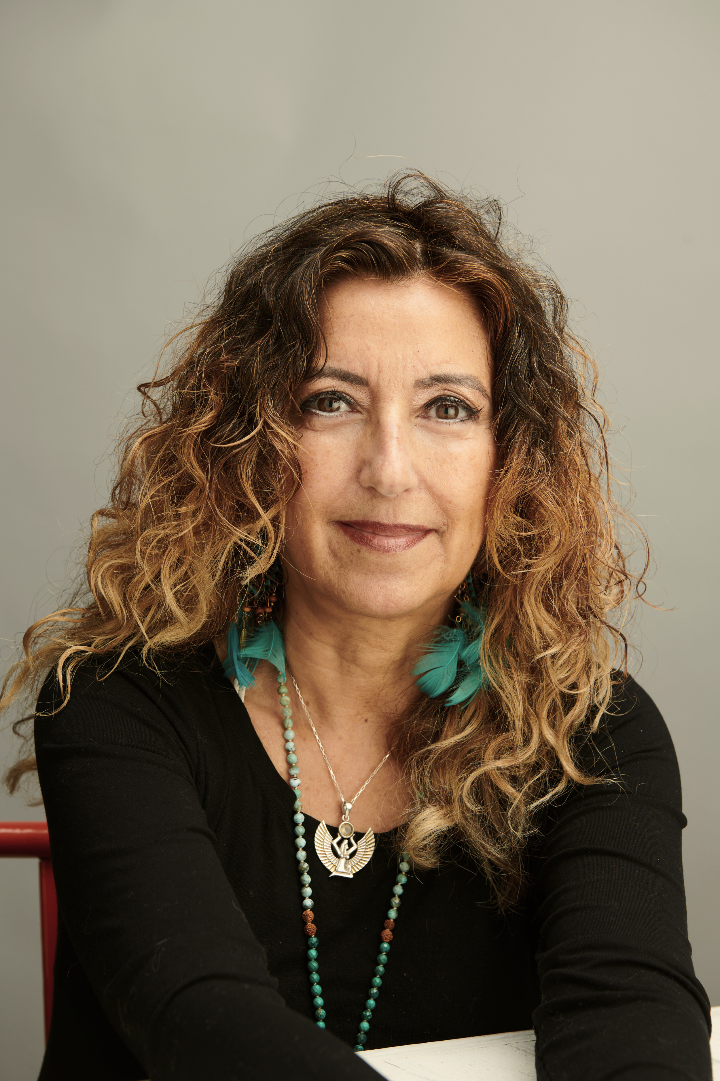 Retrato de una mujer de cabello rizado y largo, usa collar y aretes con plumas azules, lleva una prenda negra y está sentada frente a un fondo gris.