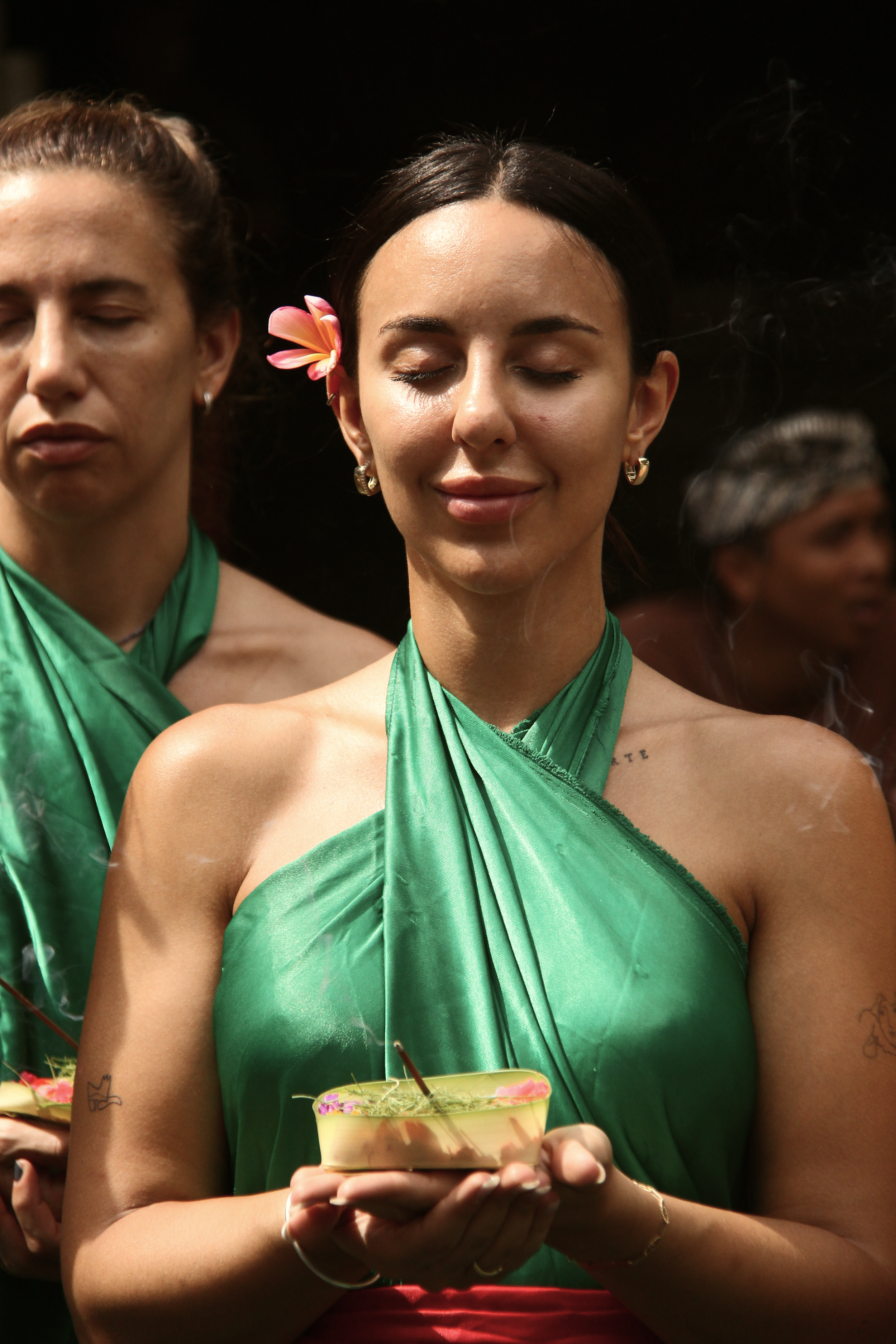 Mujer con vestido verde sosteniendo un plato con incienso, rodeada de otras personas con vestuarios similares, en un ambiente festivo o ceremonial.