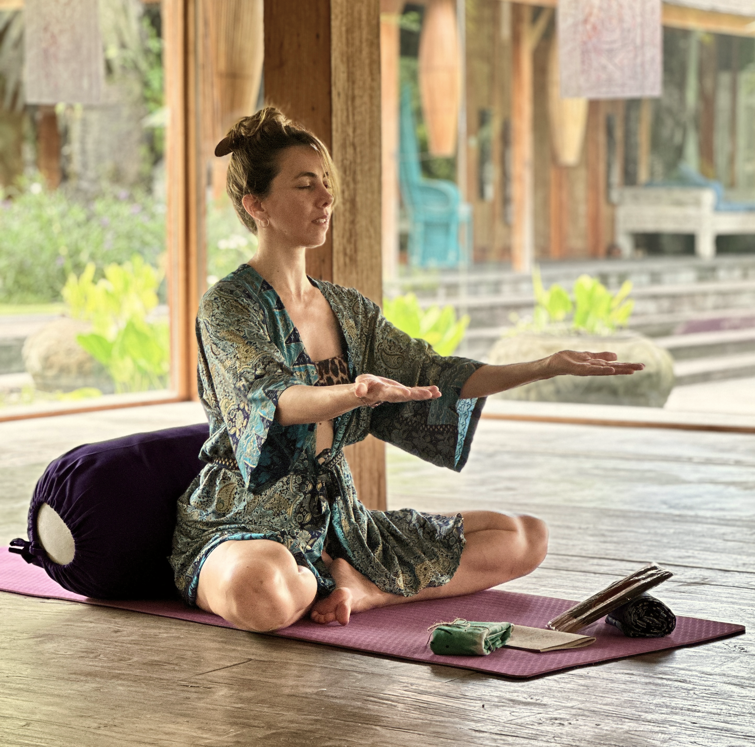 Mujer practicando yoga sentado en un tapete en un espacio de madera con ventanas grandes, rodeada de naturaleza.
