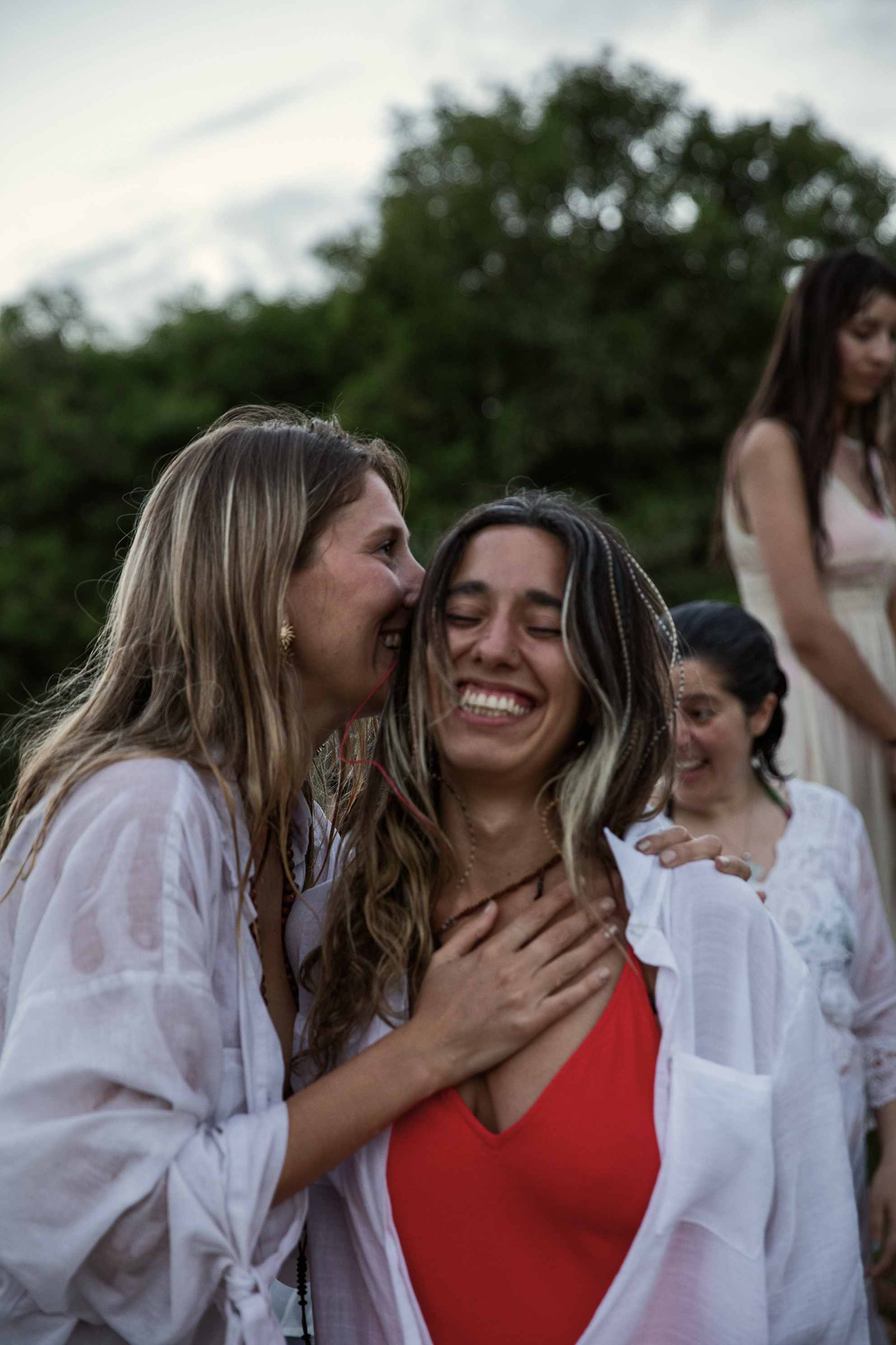 Grupo de mujeres sonriendo y abrazándose en un entorno al aire libre, con árboles y cielo de fondo, durante un evento social.
