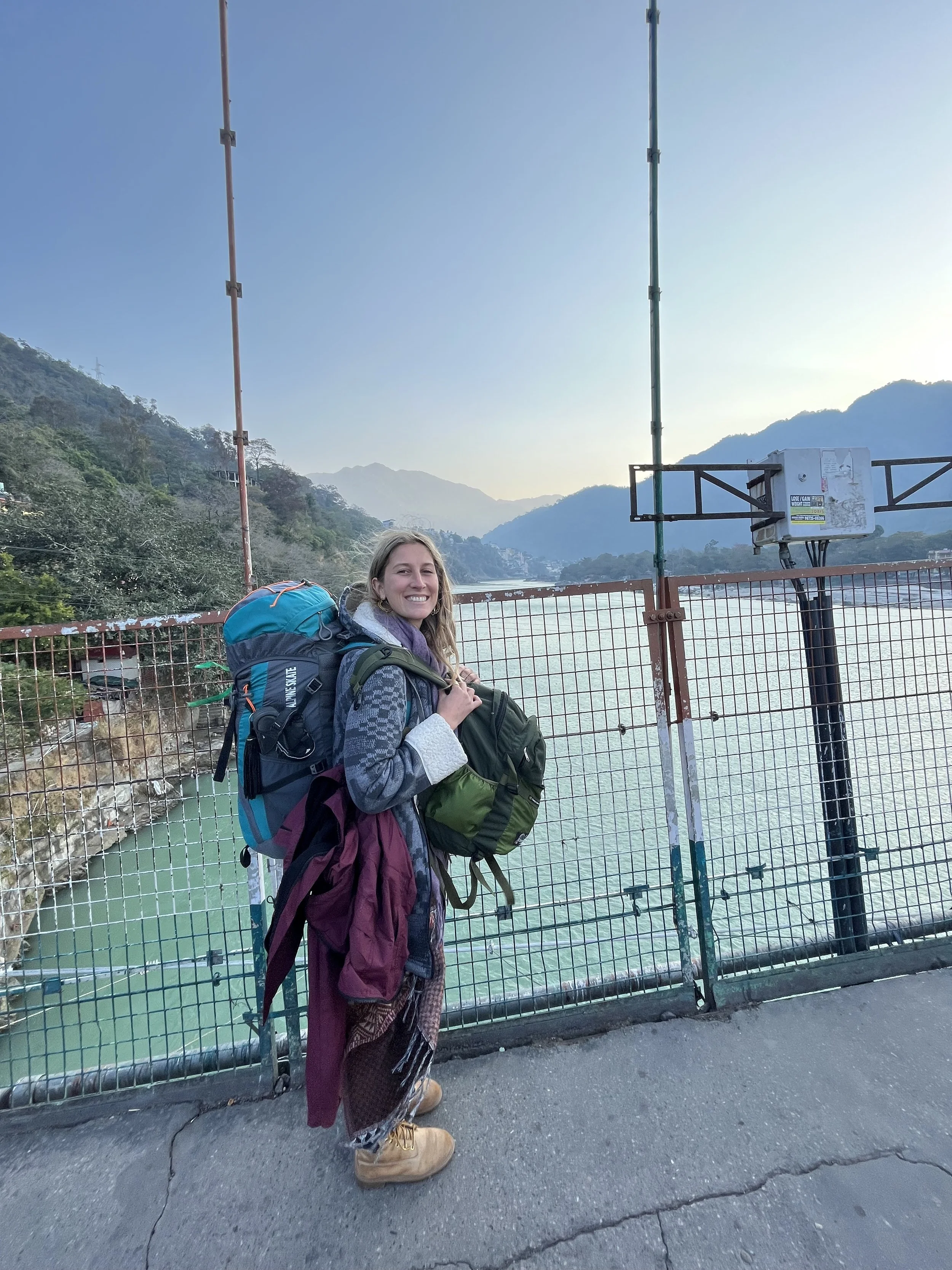 Mujer con chaqueta, mochila y bufanda de pie frente a una cerca en un puente, con un río y montañas al fondo, durante el día.