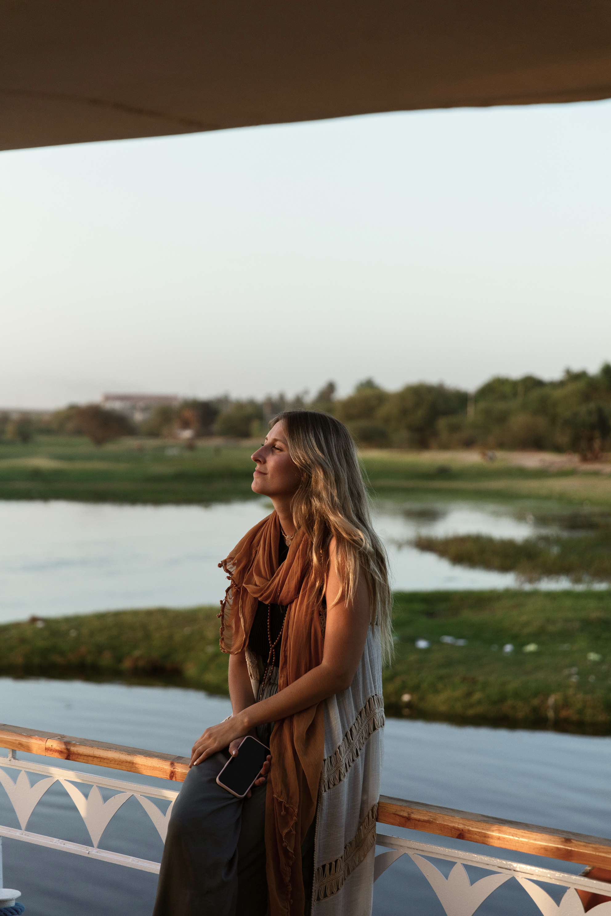 Joven mujer con cabello largo, vestimenta ligera y bufanda color marrón, de pie en una baranda junto a un río o lago, disfrutando la vista con los ojos cerrados y una expresión de paz.