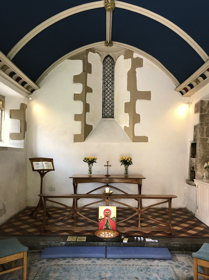 Altar de iglesia con cruces, flores y velas, dentro de un espacio con paredes blancas y ventanas pequeñas.