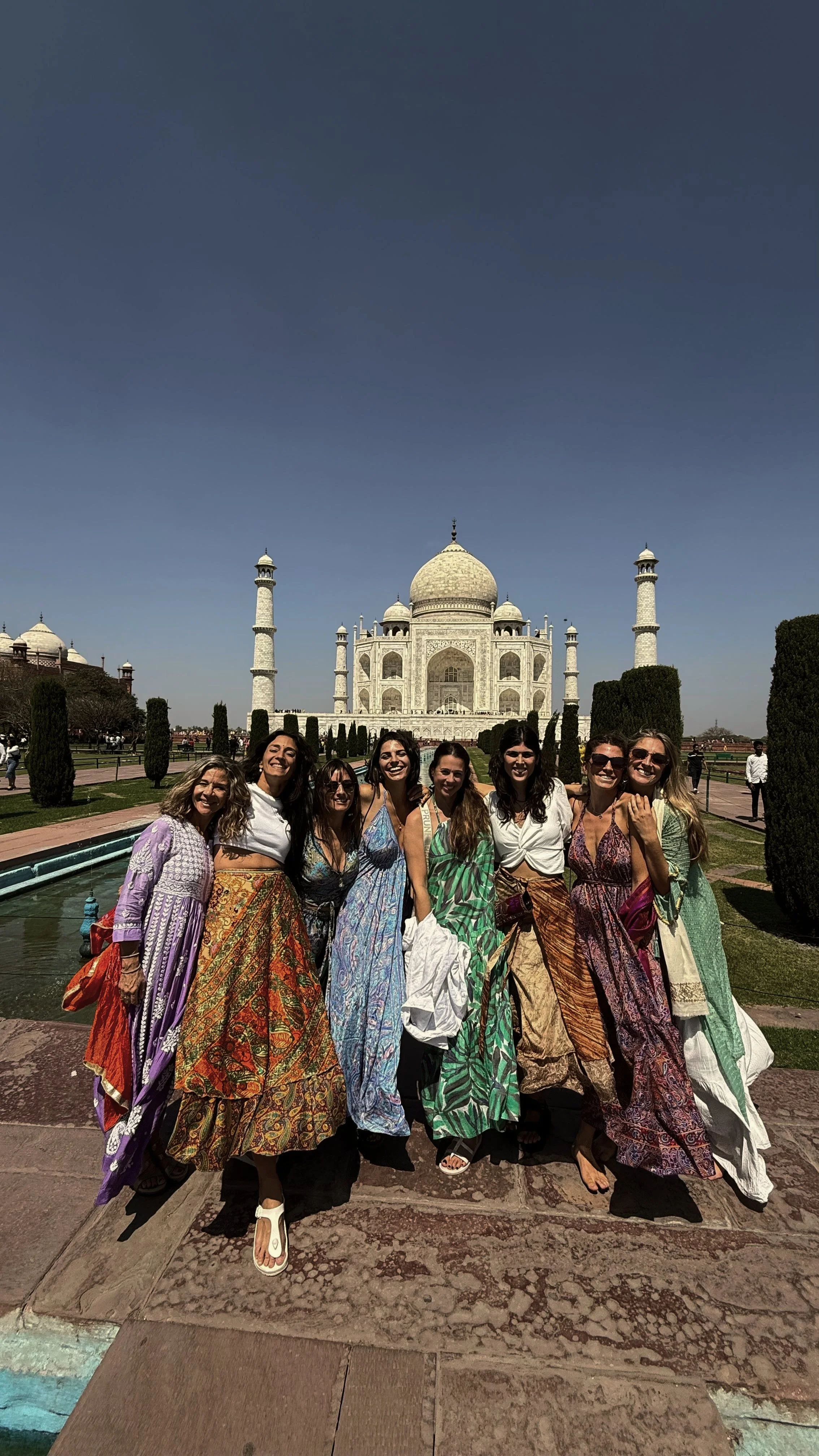 Grupo de mujeres sonriendo frente al Taj Mahal en la India, vestidas con ropas coloridas y looks veraniegos, en un día soleado.