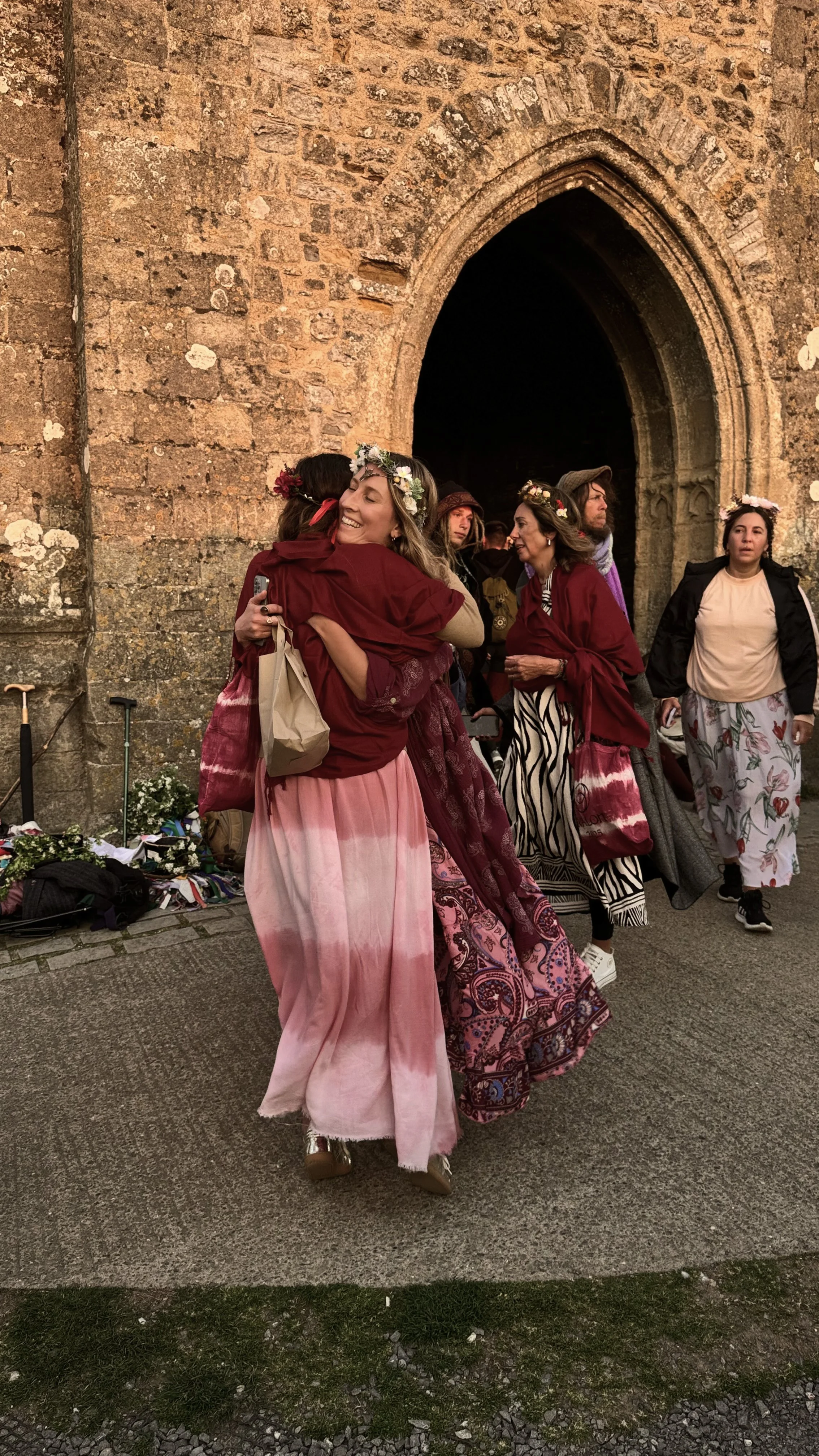 Personas en celebración con flores en la cabeza, abrazándose y sonriendo frente a una iglesia de piedra.