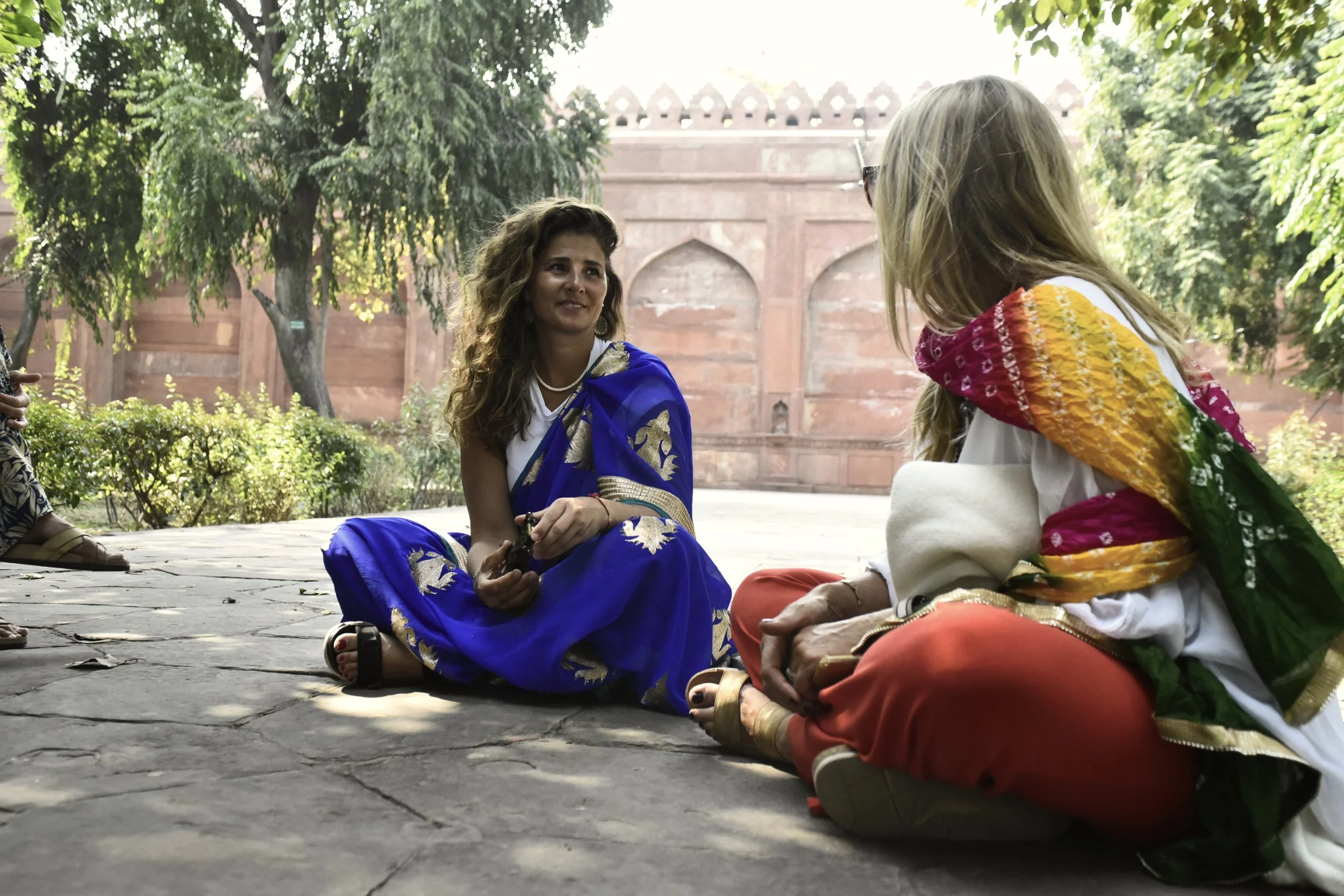 Dos mujeres sentadas en el suelo de piedra, una con un sari azul con detalles dorados y la otra con un vestido blanco y un chal multicolor, conversando en un jardín con árboles y una pared de ladrillo en el fondo.