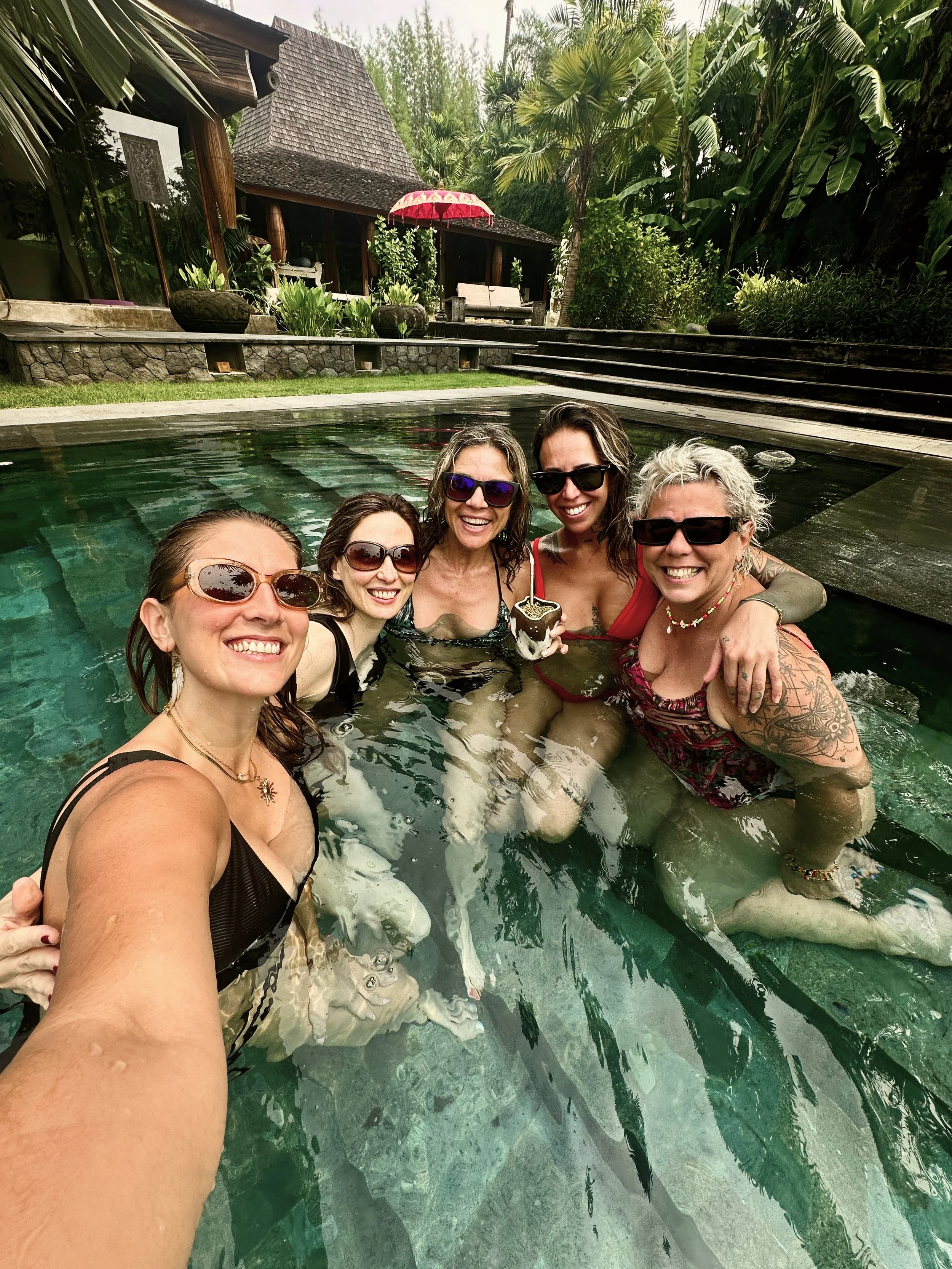 Grupo de cinco mujeres en una piscina, sonriendo y usando gafas de sol.