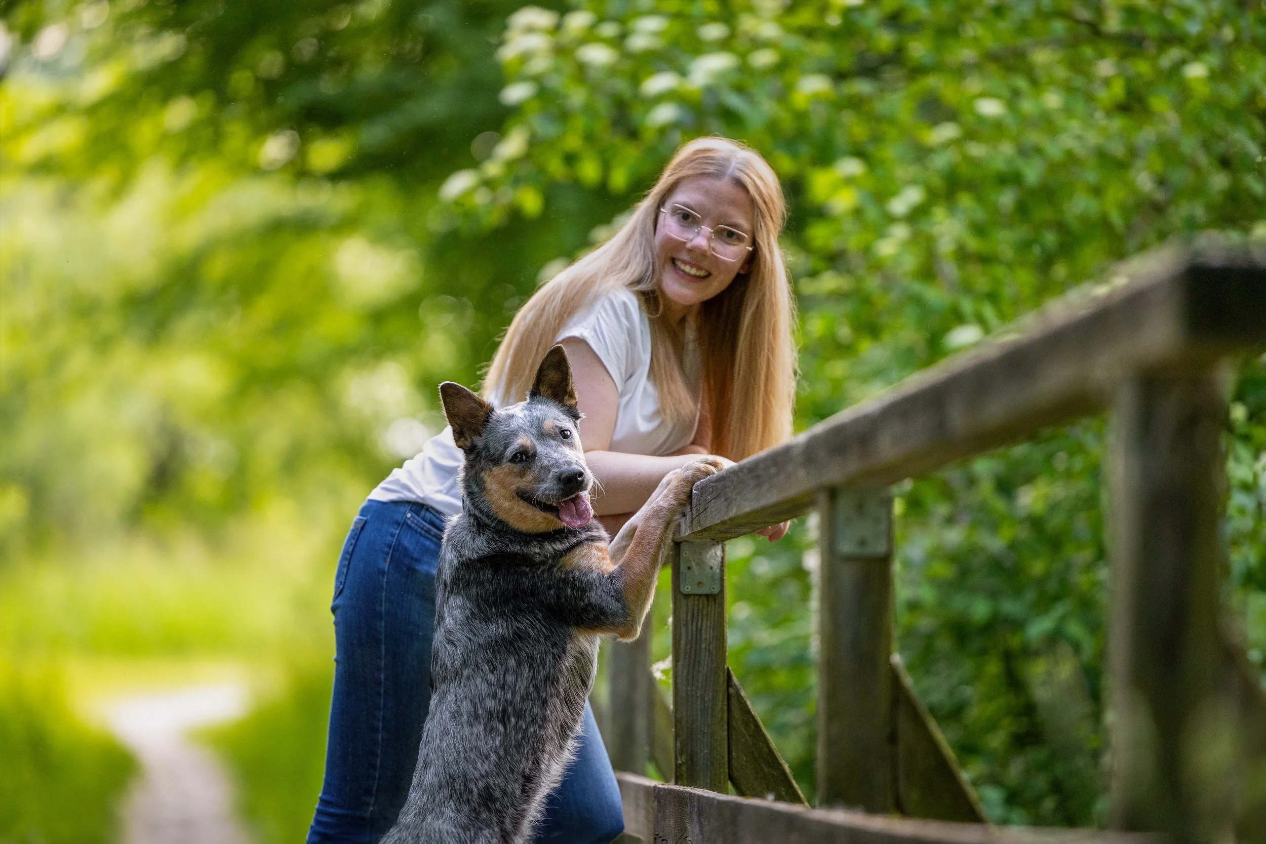 Eine lachende Frau mit langen blonden Haaren und Brille, die einen Hund an einer Pforte in einem grünen Wald am Tag hält.