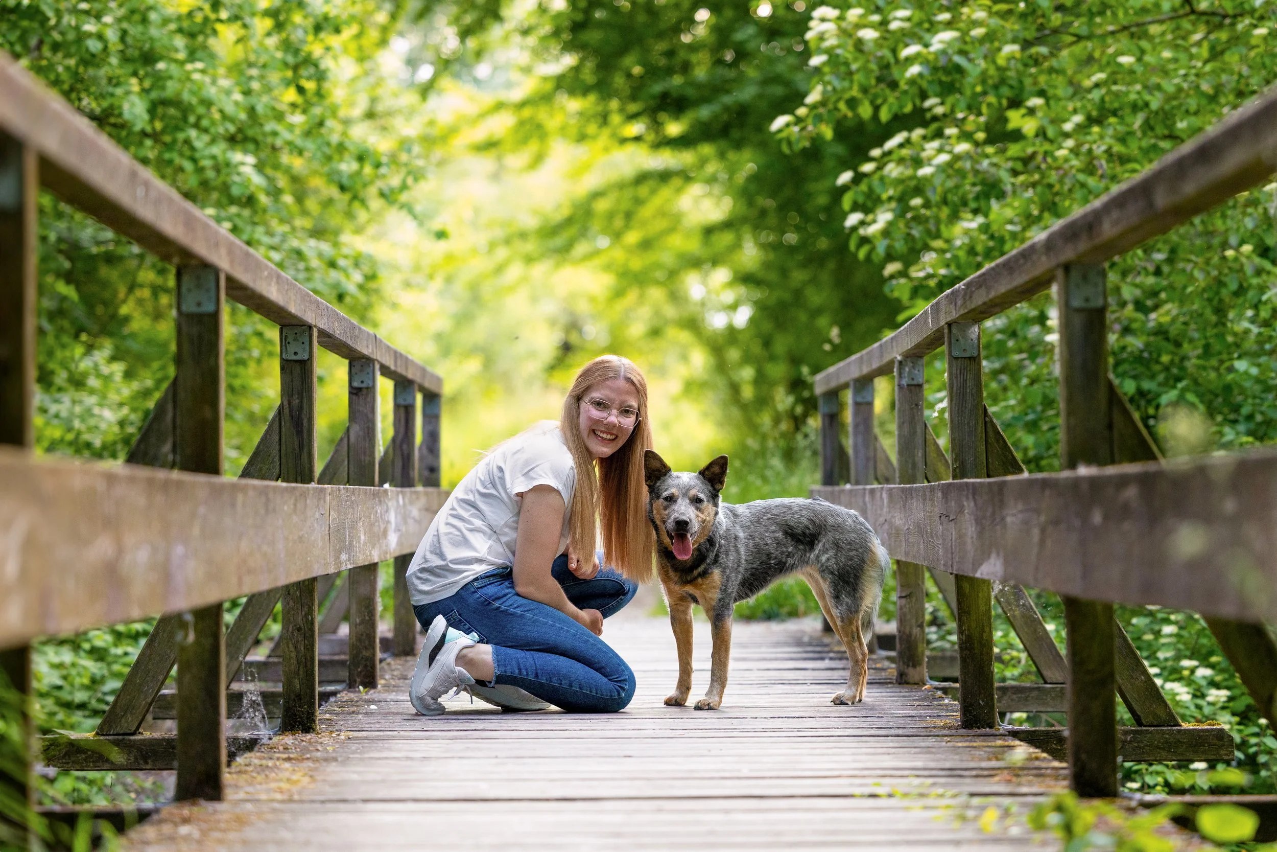 Eine lachende Frau kniet neben einem grauen und schwarzen Hund auf einer Holzbrücke in einer grünen, bewaldeten Umgebung.