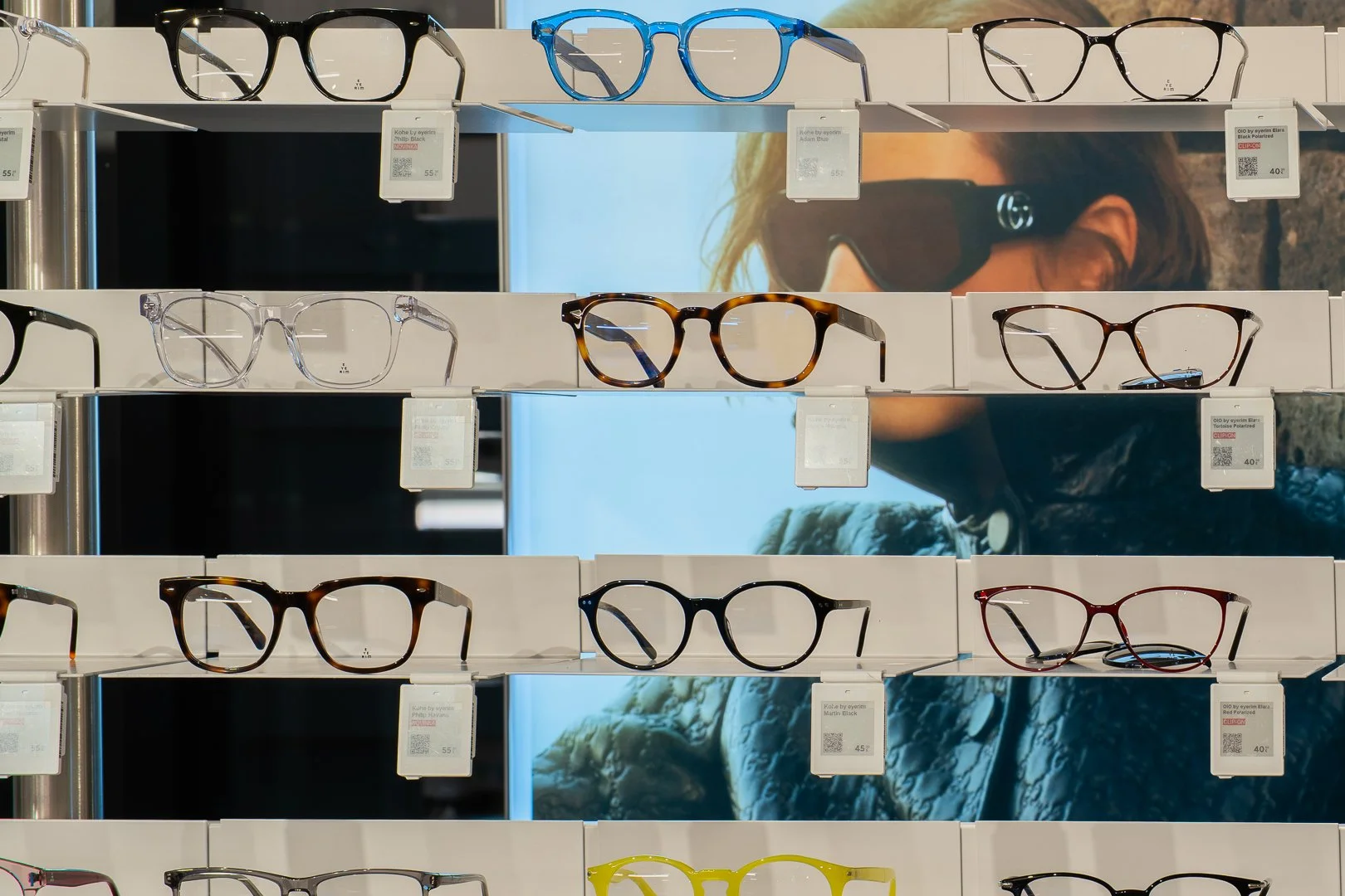 Dioptrické okuliare vystavené na polici v predajni, s plagátom osoby so slnečnými okuliarmi v pozadí.