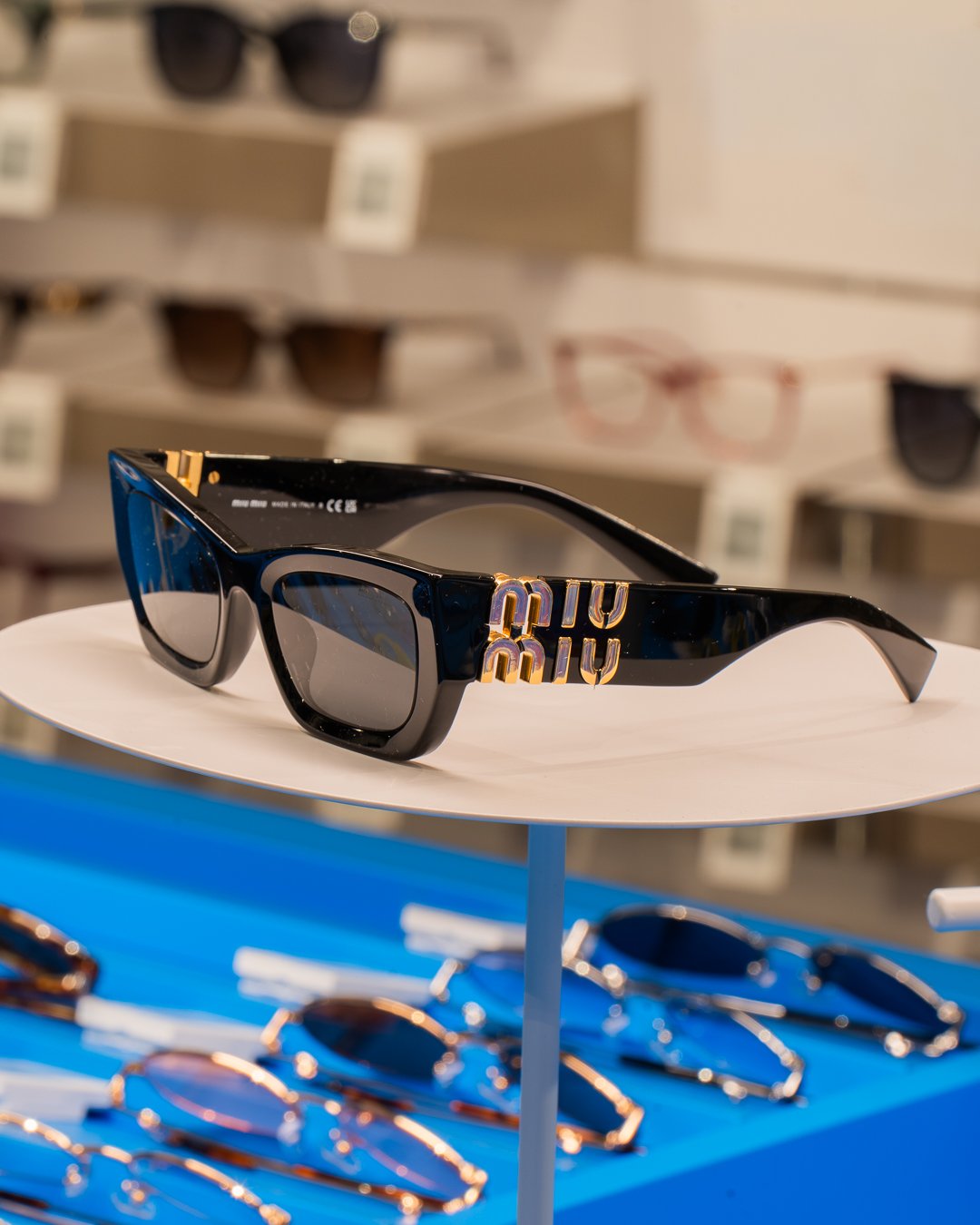Slnečné okuliare Miu Miu s čiernym rámom a zlatými detailmi vystavené na bielom stojane v predajni.
