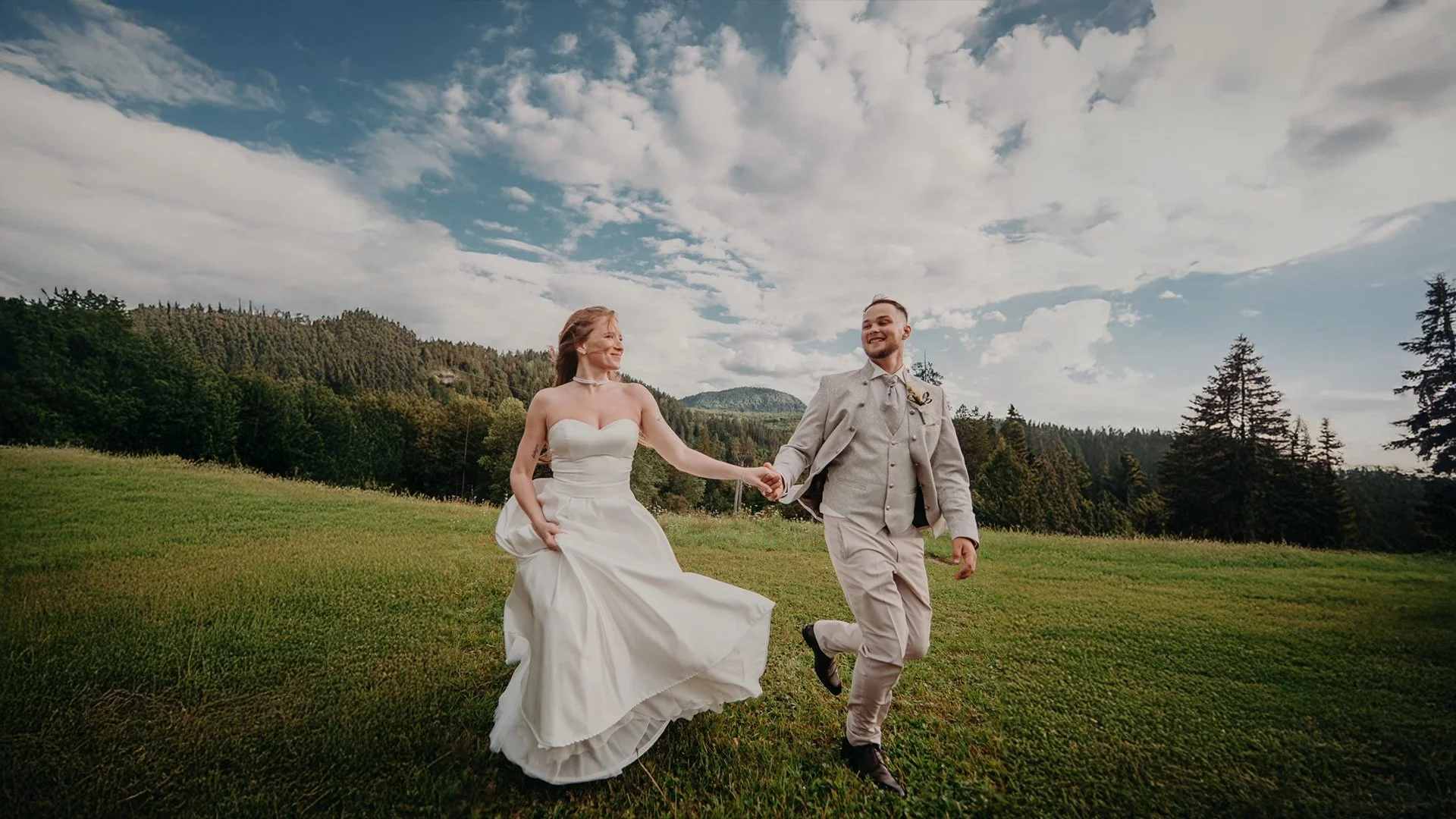 Novomanželia bežia ruka v ruke po lúke so stromami a kopcami v pozadí.
