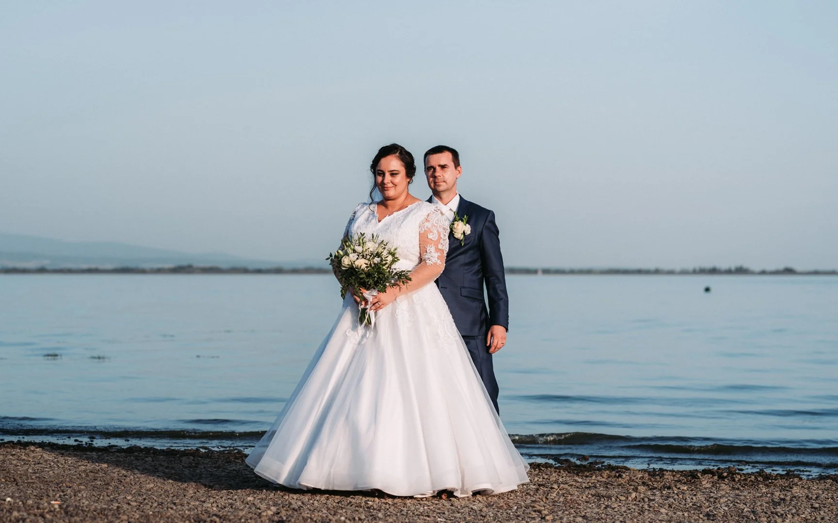 A beige ceramic vase with a narrow opening, holding a dried flower, placed on a wooden surface with sunlight casting a shadow on the wall behind.
