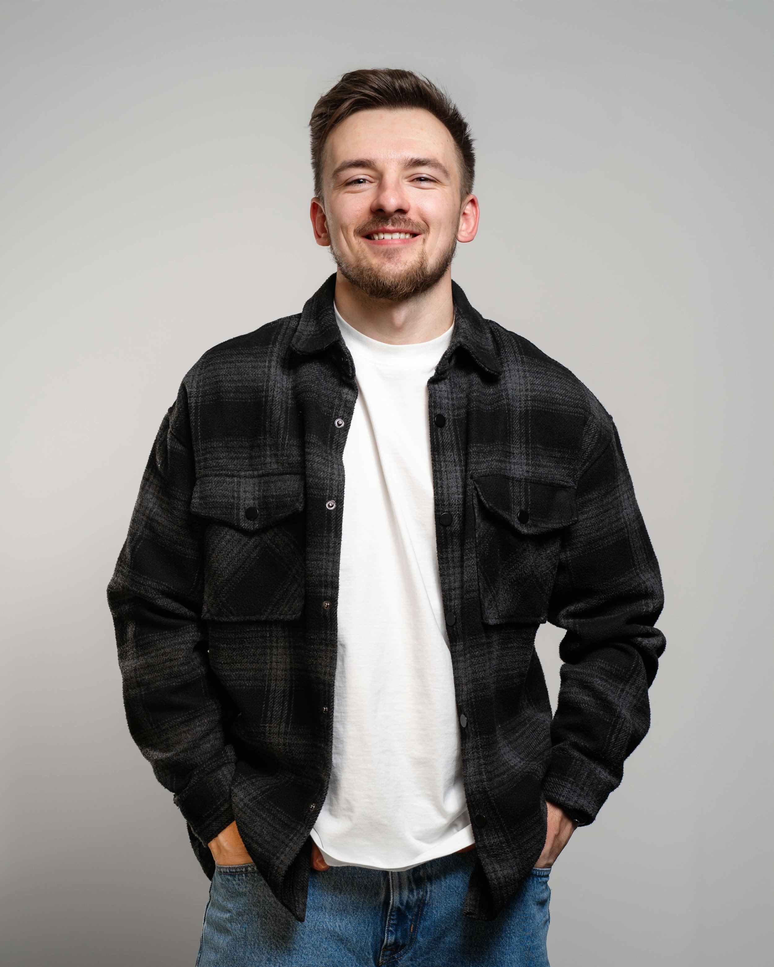 Portrét mladého muža s bradou v kockovanej košeli, usmieva sa na svetlom pozadí.