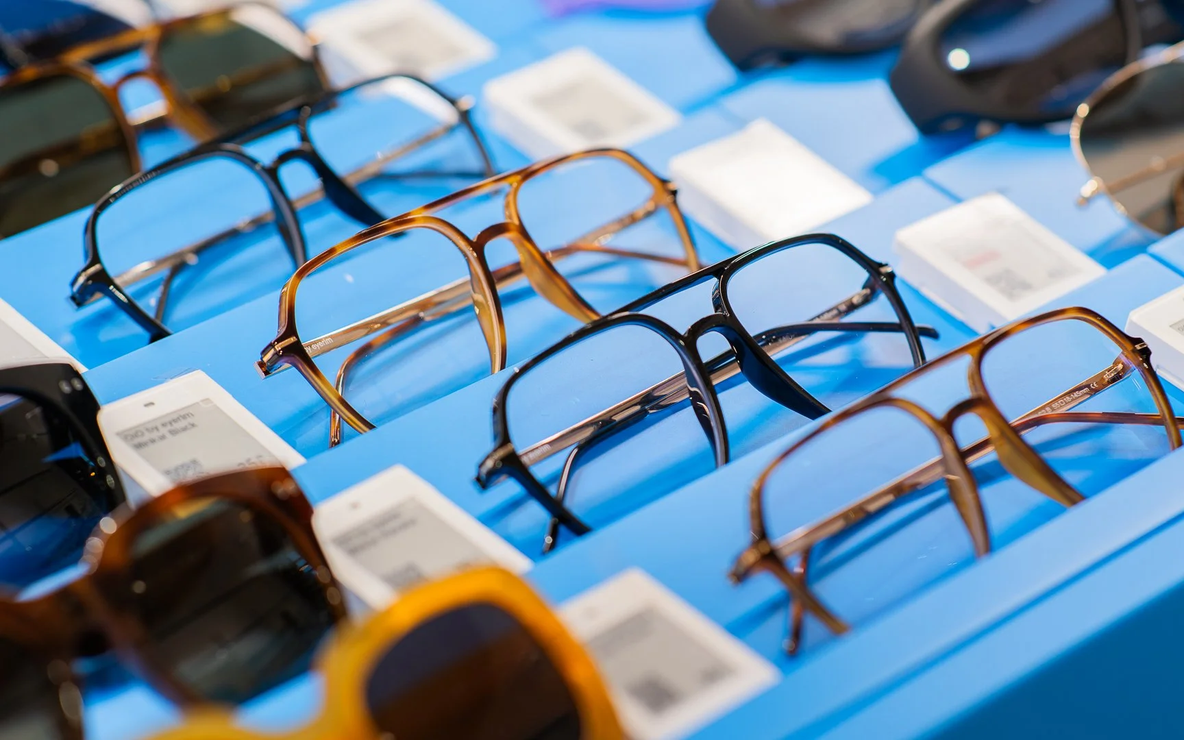 Display of various eyeglasses and sunglasses on a blue table at an optical store.