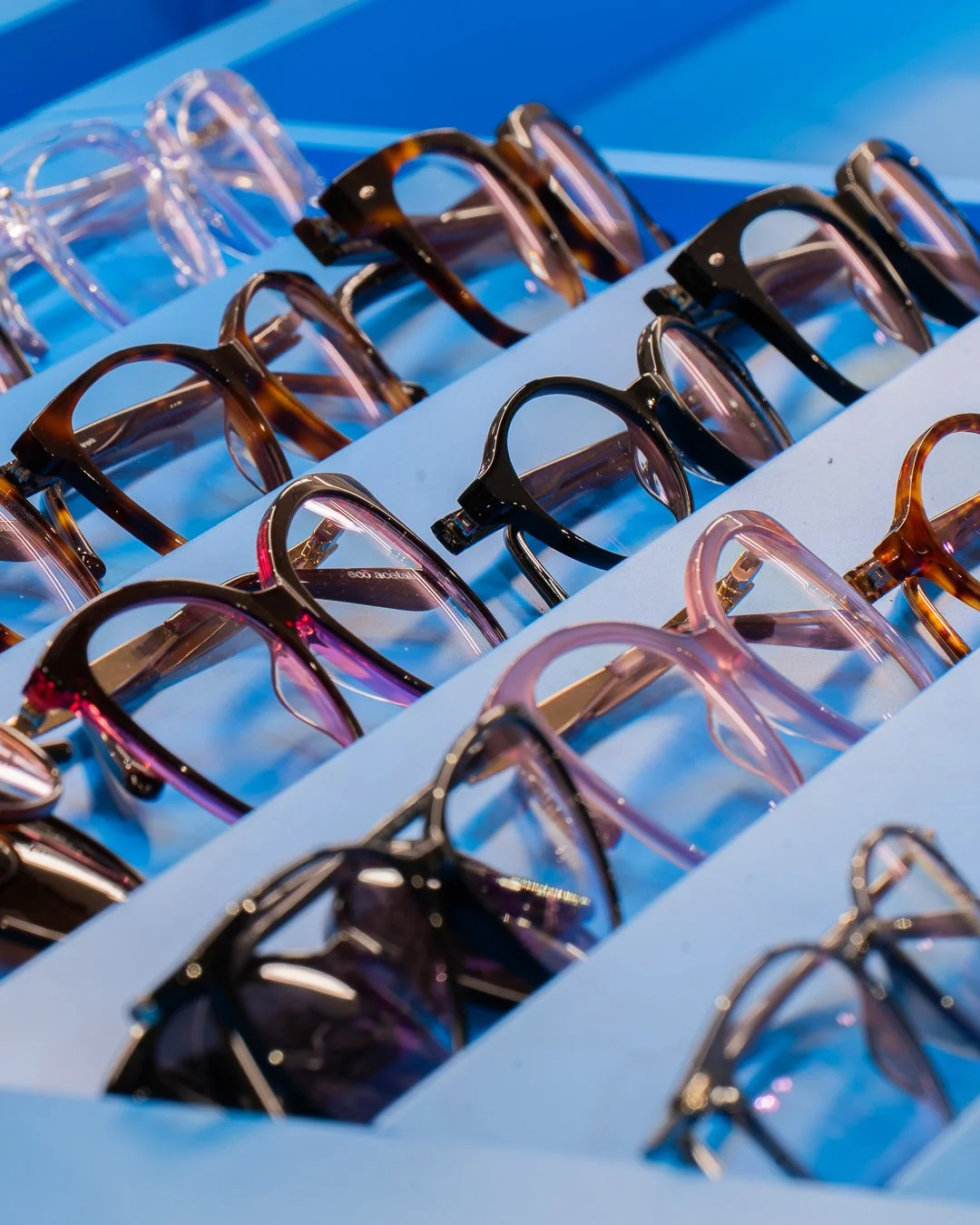 Rôzne slnečné okuliare vystavené v radoch na svetlomodrom stojane s modrým pozadím.