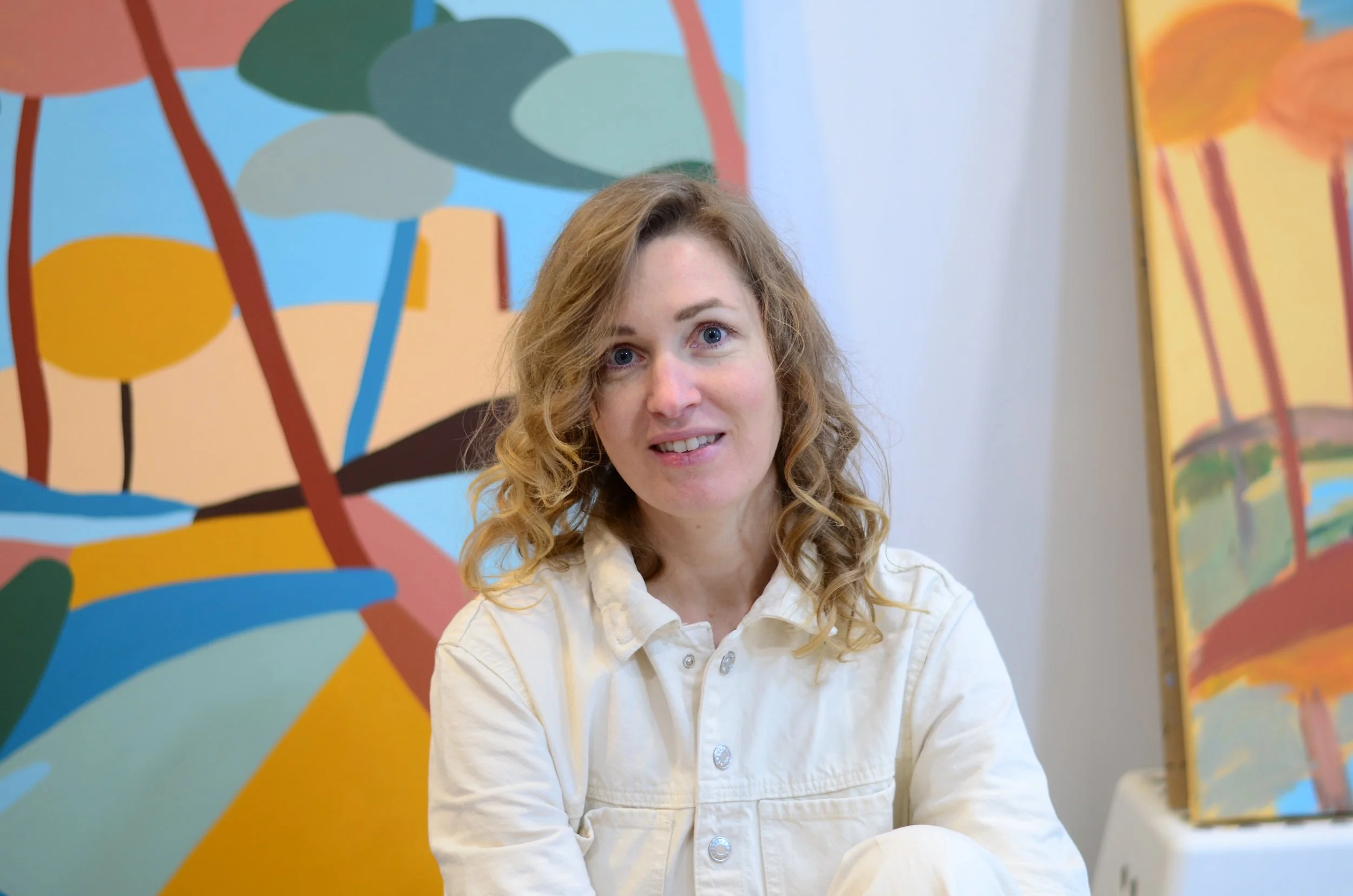 Une femme aux cheveux bouclés, portant une veste claire, assise devant une peinture abstraite colorée avec des formes organiques et des arbres stylisés.