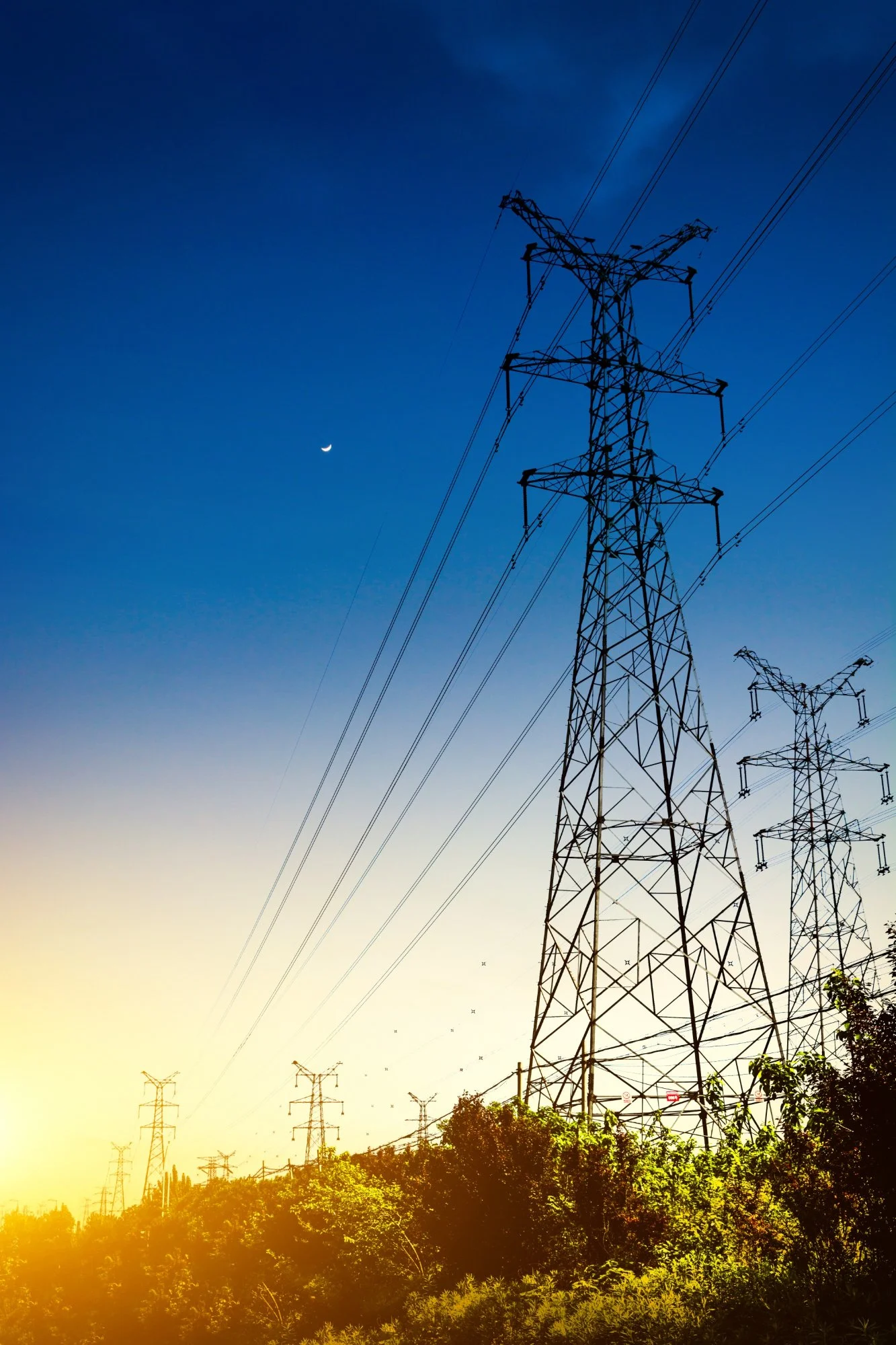 A row of electric power transmission towers against a sunset sky with a crescent moon.