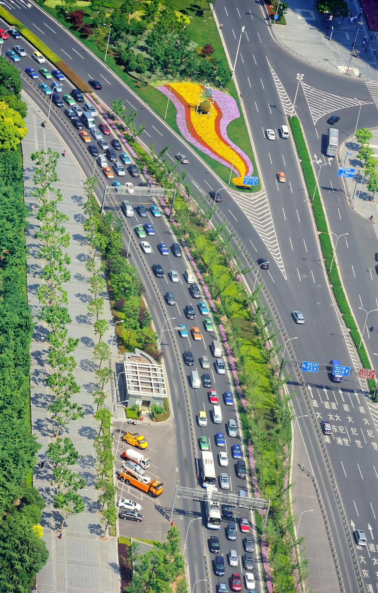 An aerial view of a city street with multiple lanes of traffic, a green landscaped median with colorful flowers in a pattern, a sidewalk with trees, and surrounding buildings.