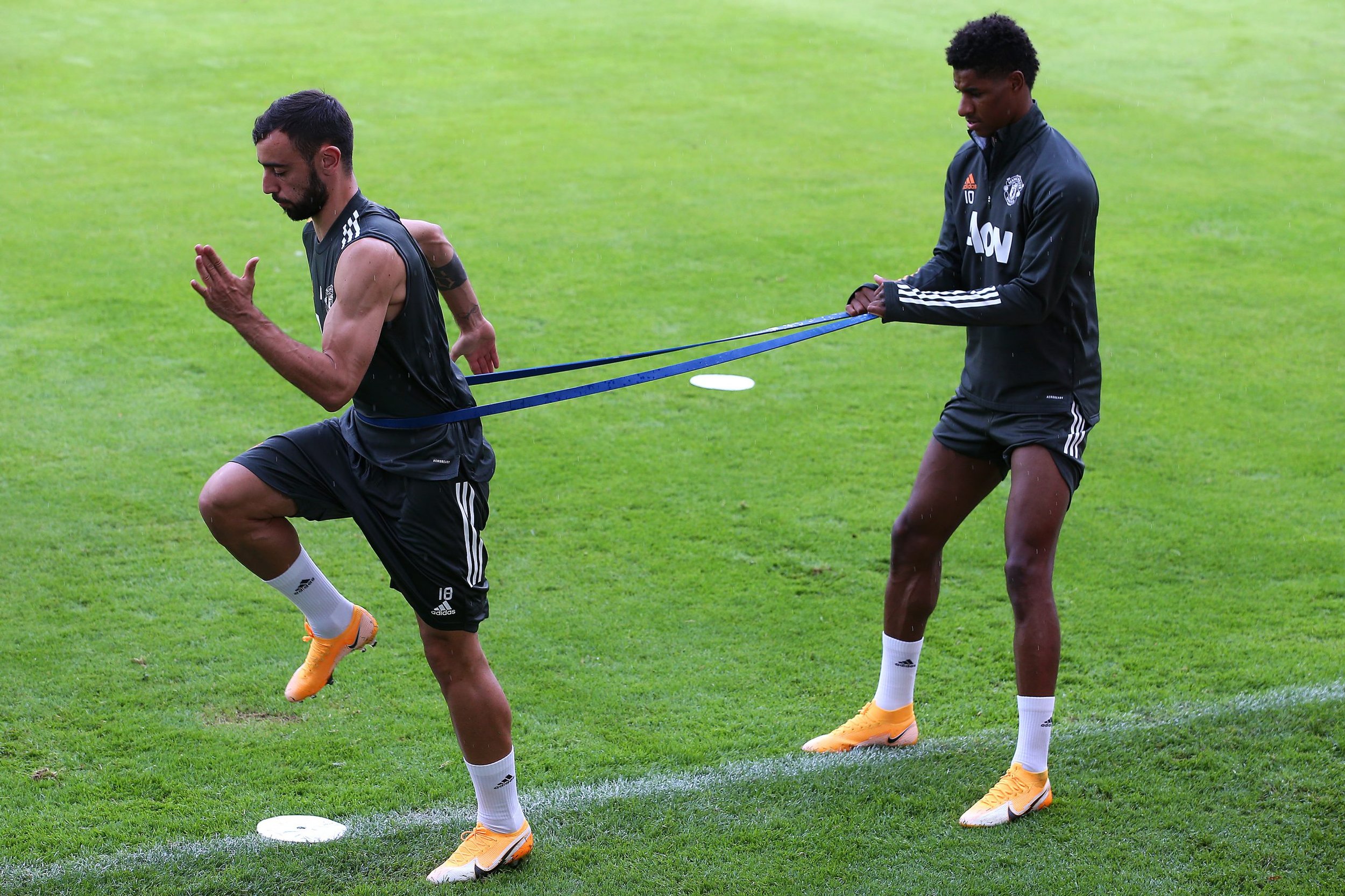 Zwei Fußballspieler auf einem Rasenplatz, einer führt ein Lauftraining durch, der andere zieht ihn mit einem Widerstandsband. Beide tragen schwarze Sportkleidung mit Adidas-Logo und orange Schuhe.
