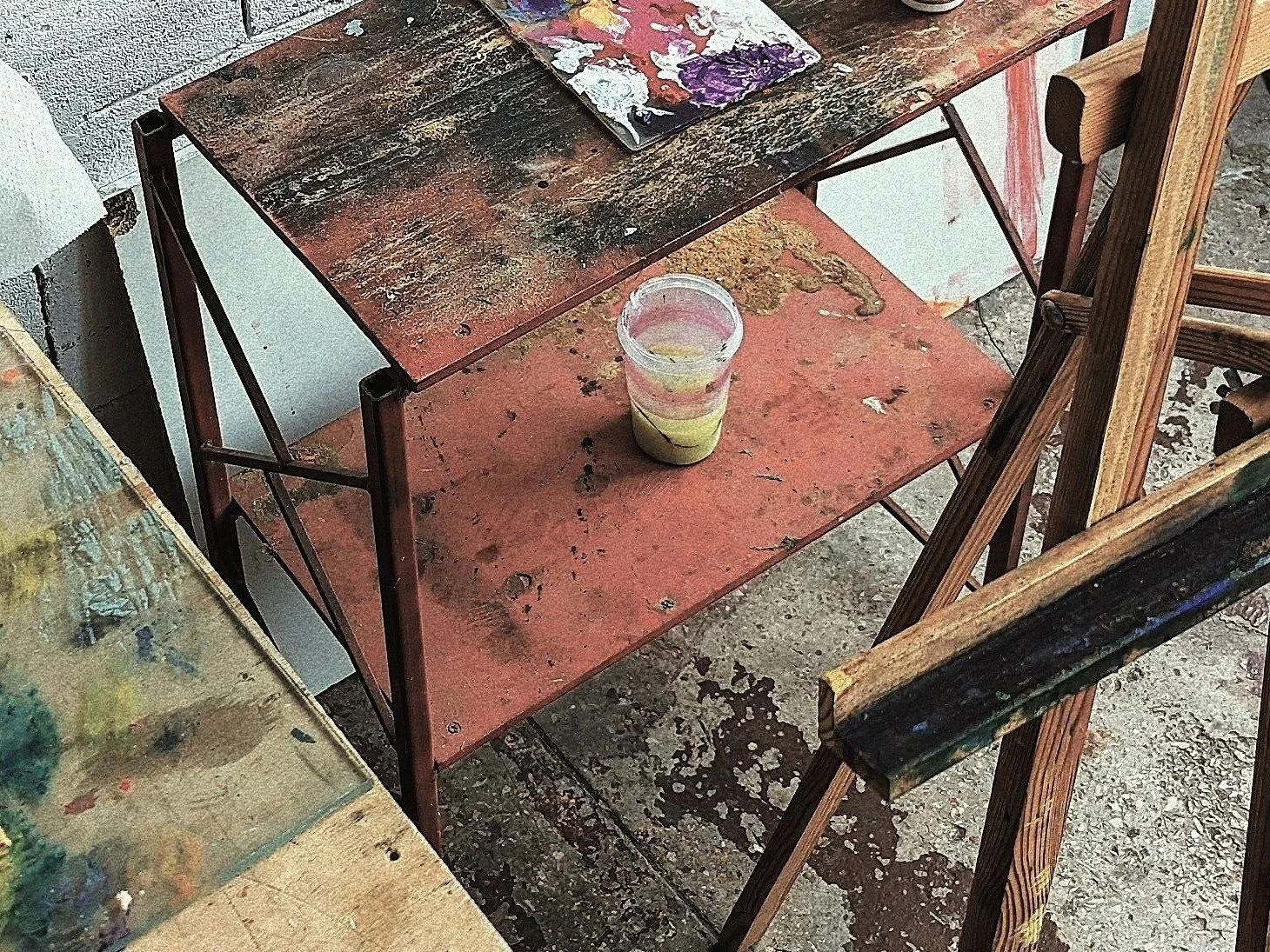 Paint-stained art table with a glass of yellowish paint, surrounded by other paint-stained surfaces and an easel.