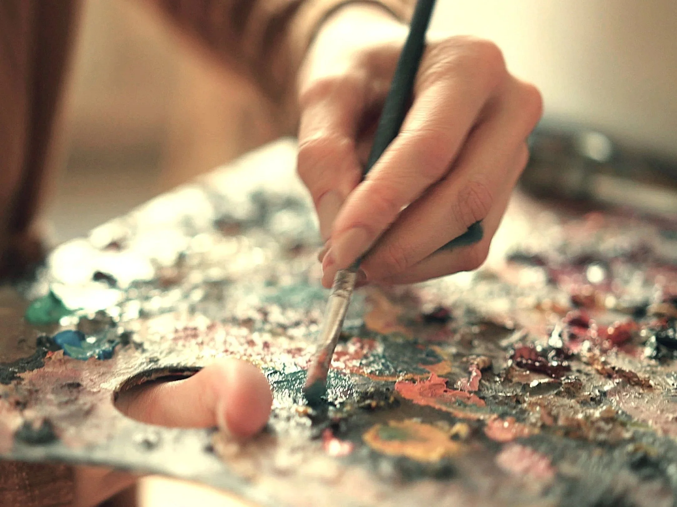 Close-up of a person’s hand using a paintbrush to paint on a multicolored palette or surface with paint.