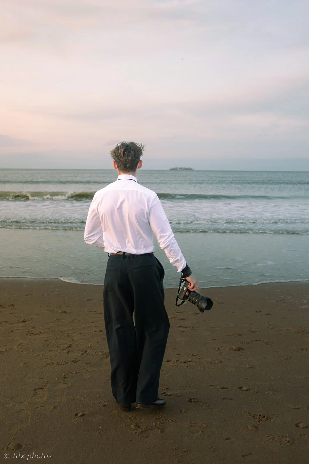 Un homme en pantalon large et chemise blanche tient un appareil photo au bord de la mer, regardant l'horizon, avec un navire en arrière-plan dans le ciel.