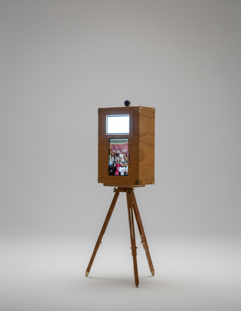 Cabine photo en bois avec écran et imprimante, sur un trépied en bois, dans un espace blanc.