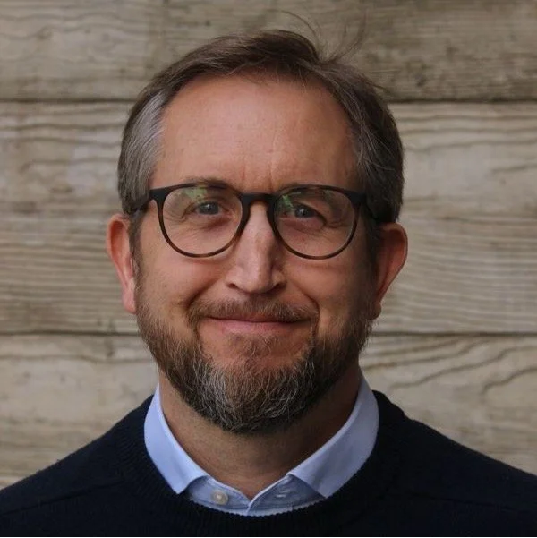 This is a picture of the founder of Baker Aventine, Luc Salmon, a middle-aged man with glasses, a beard, and gray hair smiling at the camera, standing in front of a wooden wall.