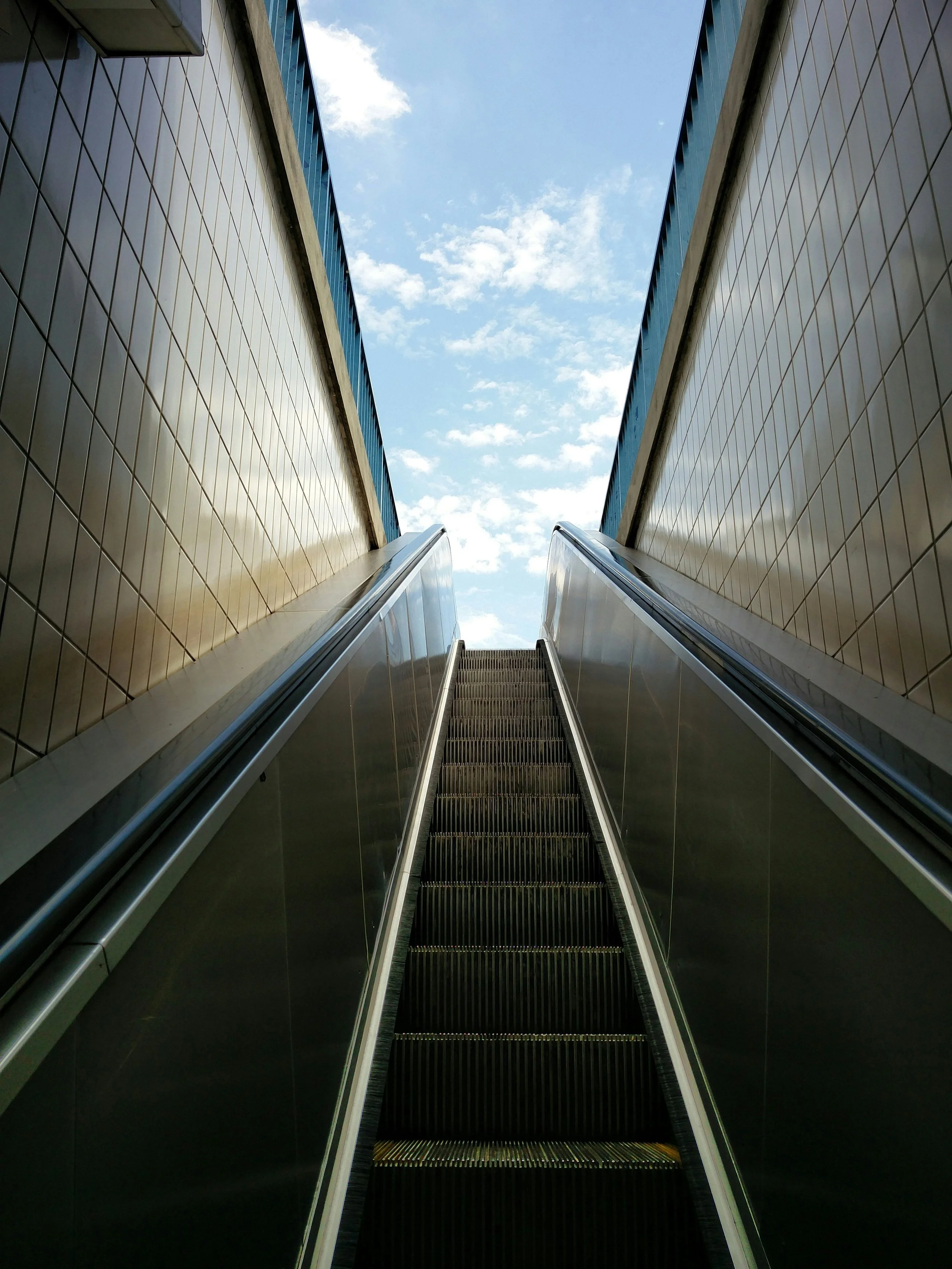View of an upward escalator between two tall buildings with beige tiled walls, leading towards a cloudy blue sky. This picture represents the commitment BakerAventine makes to support Social Mobility projects.